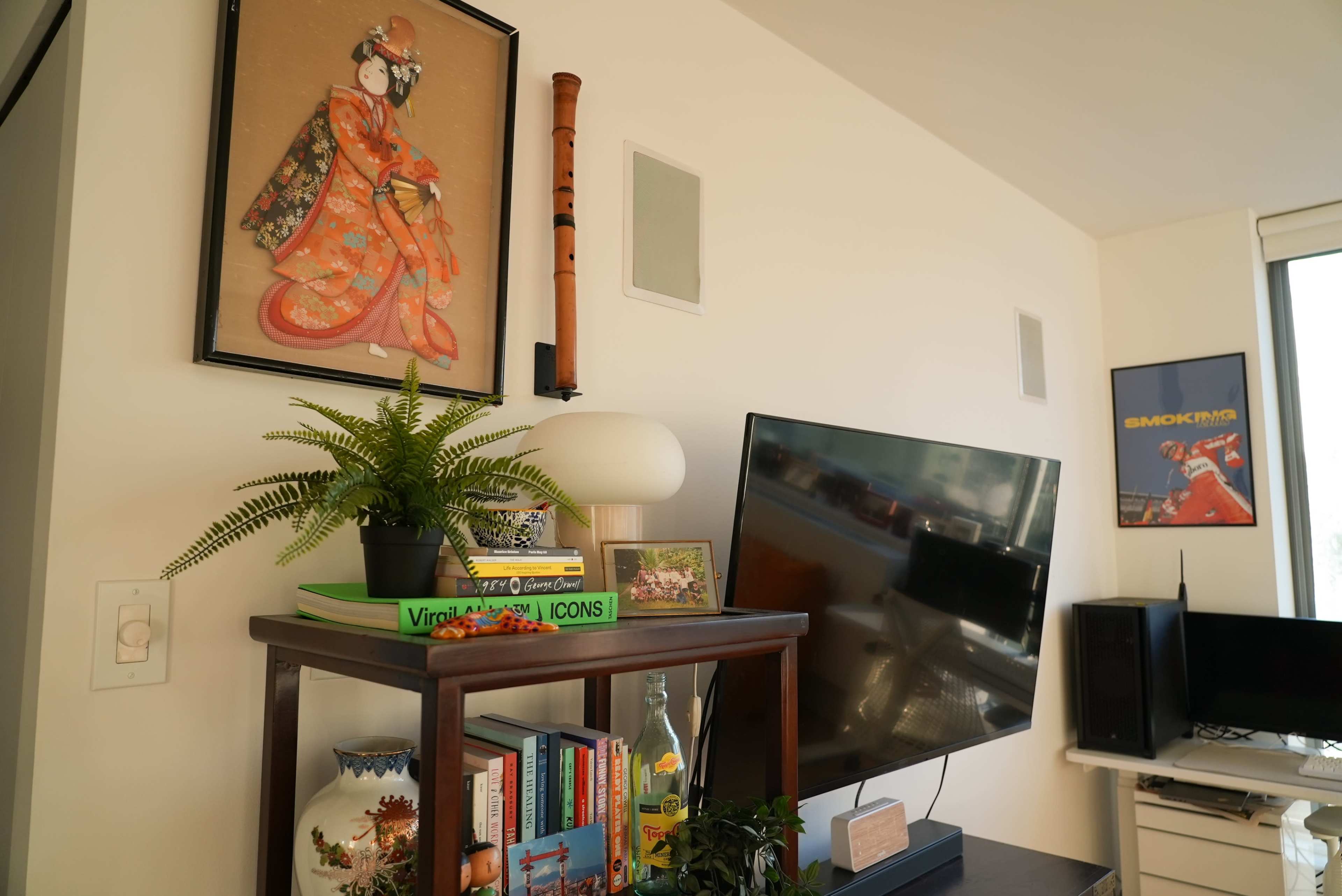 The scene features a wall adorned with a framed artwork of a geisha, a television on a wooden stand, and various decorative plants and books beside it.