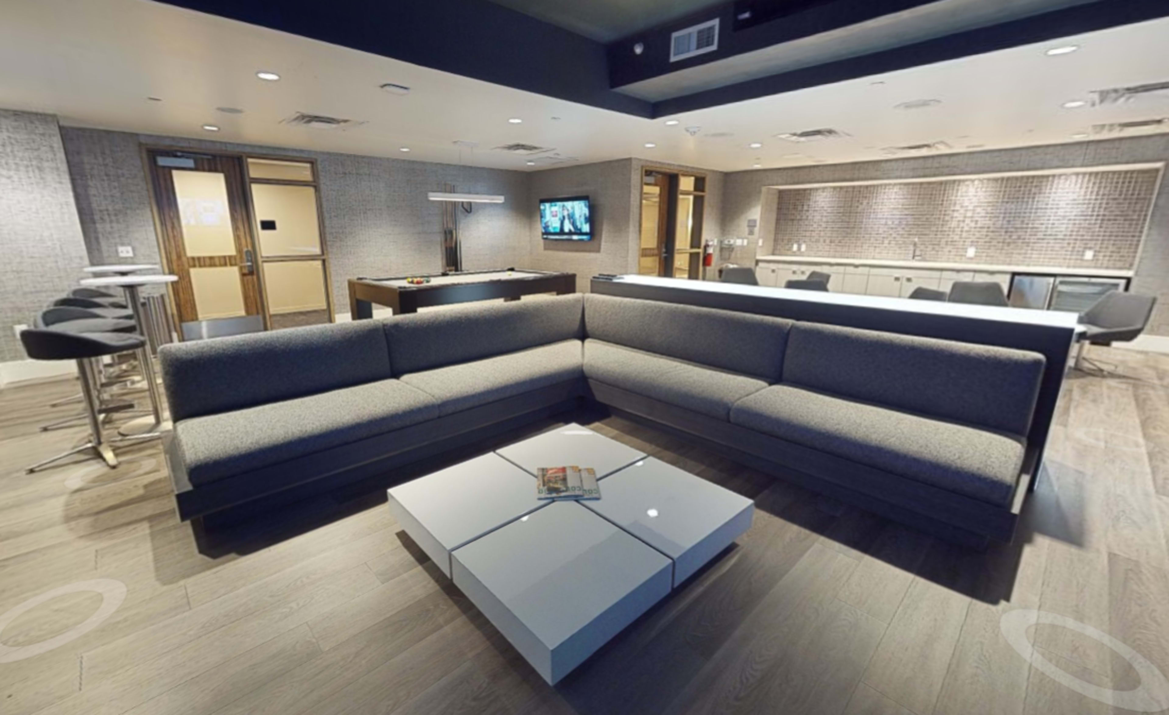 The image shows a modern lounge area featuring a gray sectional sofa, a square coffee table, and a flat-screen TV on the wall, with an open kitchen space visible in the background.