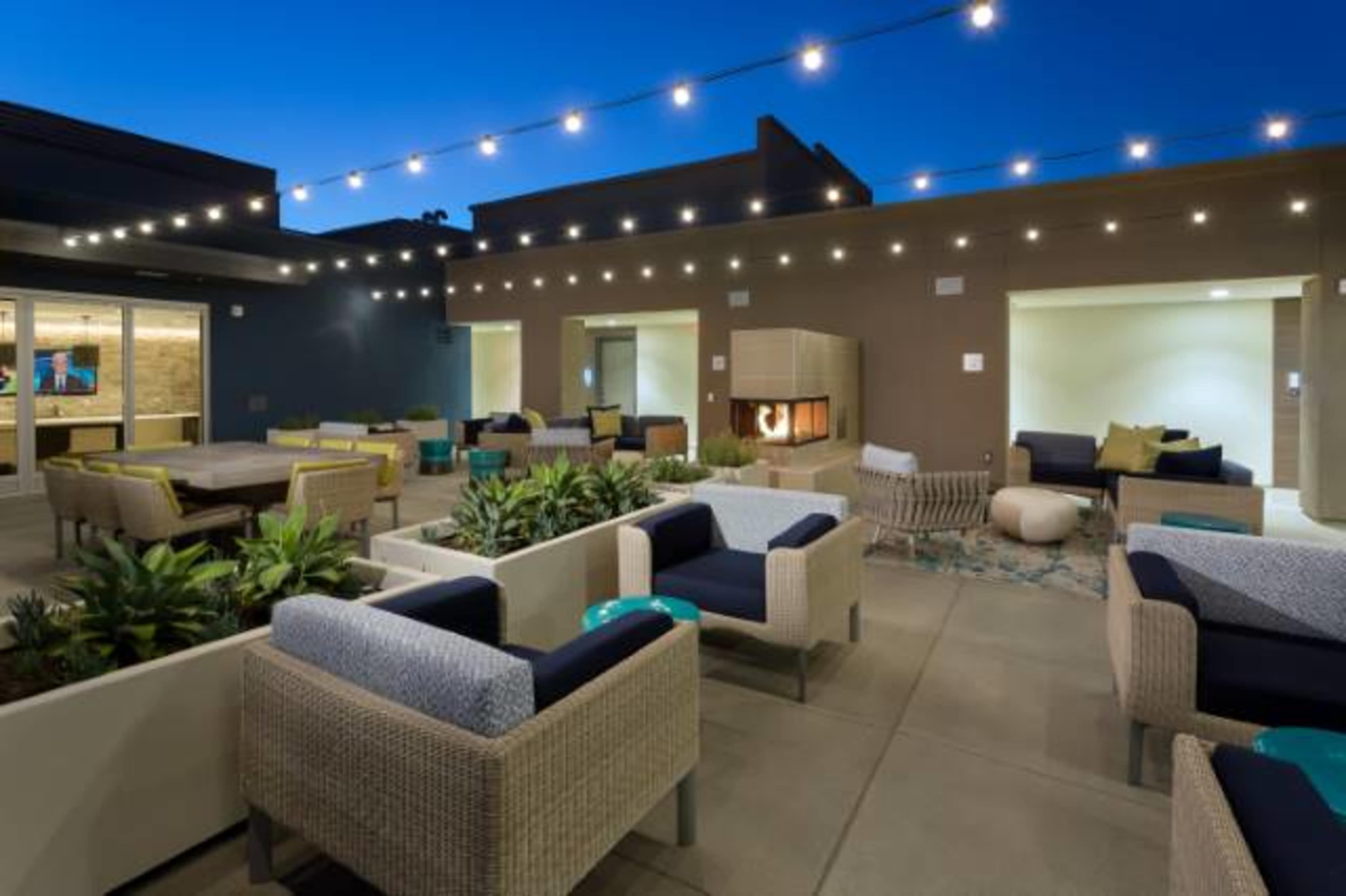 A modern outdoor lounge area with seating, fire features, and string lights illuminating the space at dusk.