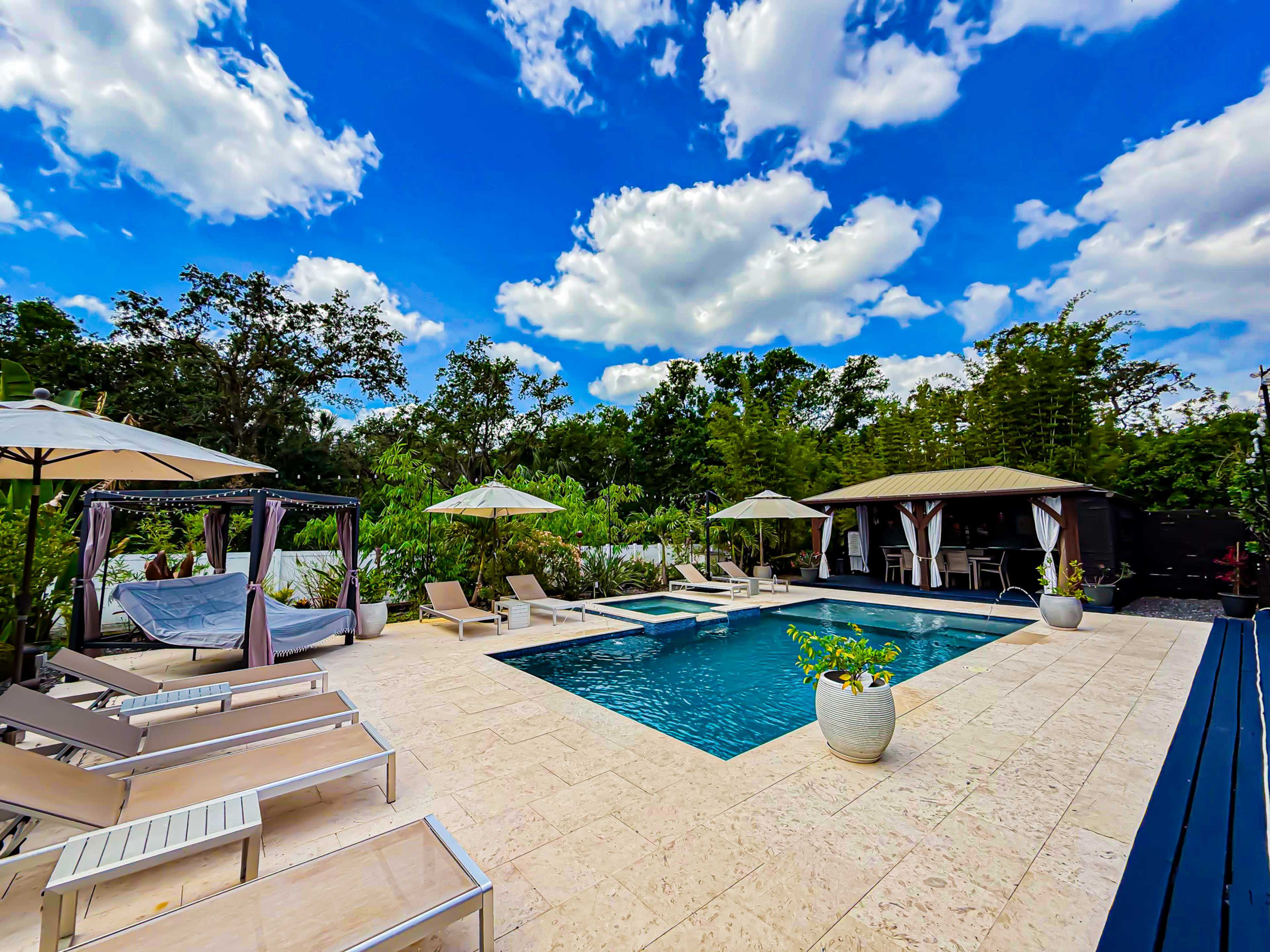A rectangular pool is surrounded by lounge chairs, umbrellas, and a gazebo, with lush greenery and a building in the background under a partly cloudy sky.