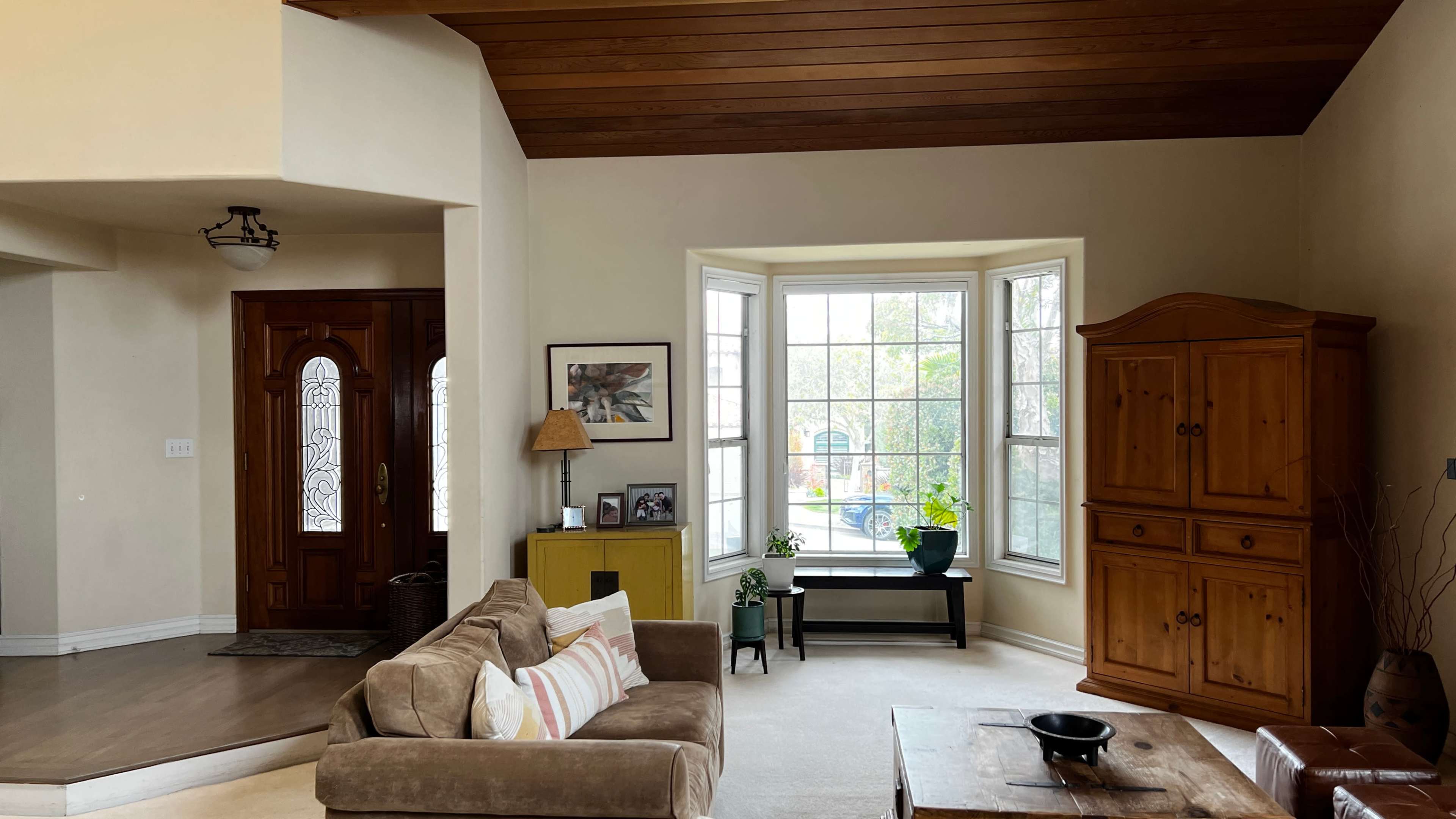 A living room with a large window, wooden ceiling beams, a beige sofa, a wooden cabinet, and a small table.