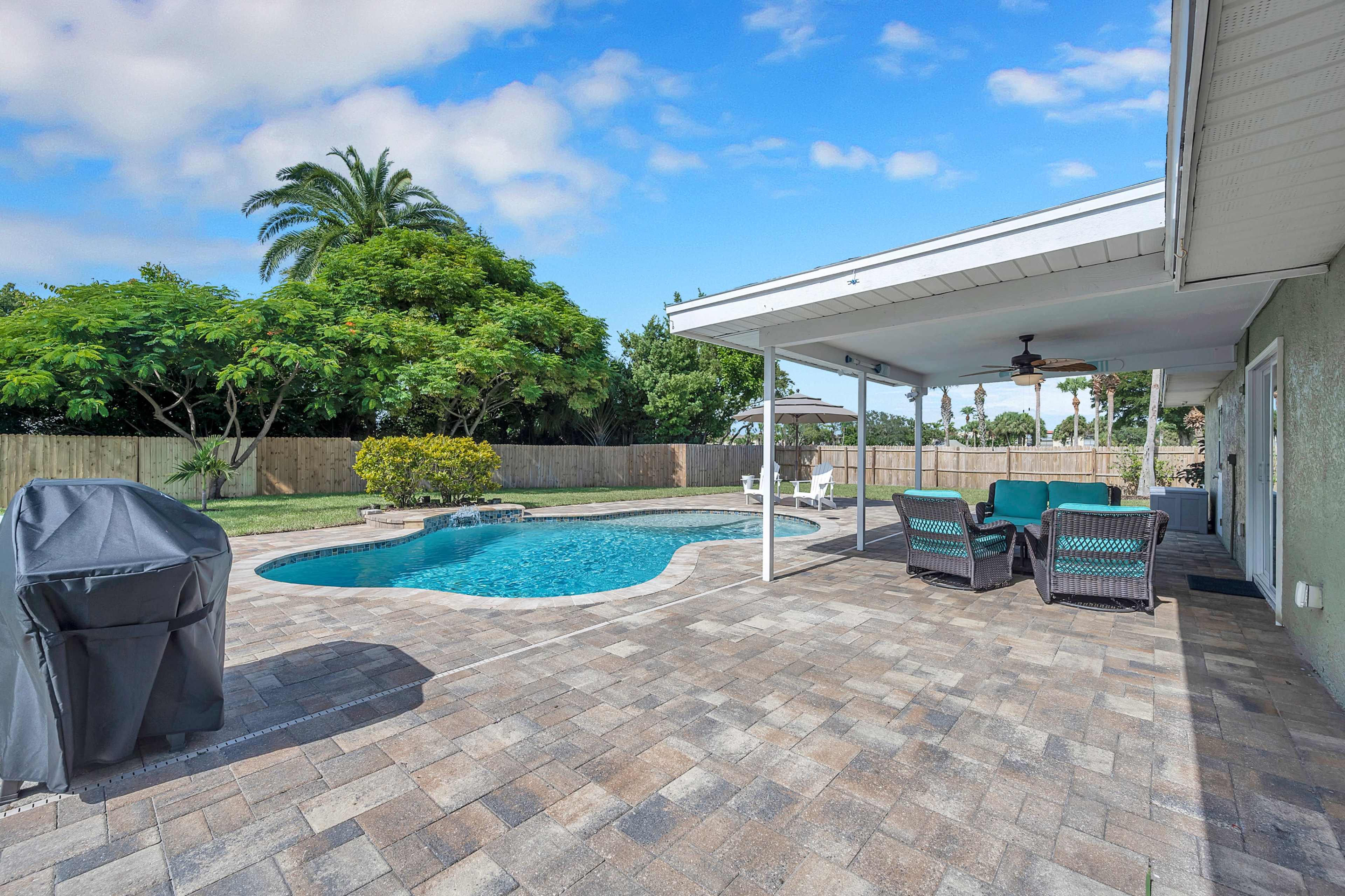 The image depicts a backyard with a swimming pool surrounded by stone paving, outdoor seating, and a grill under a covered patio, bordered by greenery and a wooden fence.