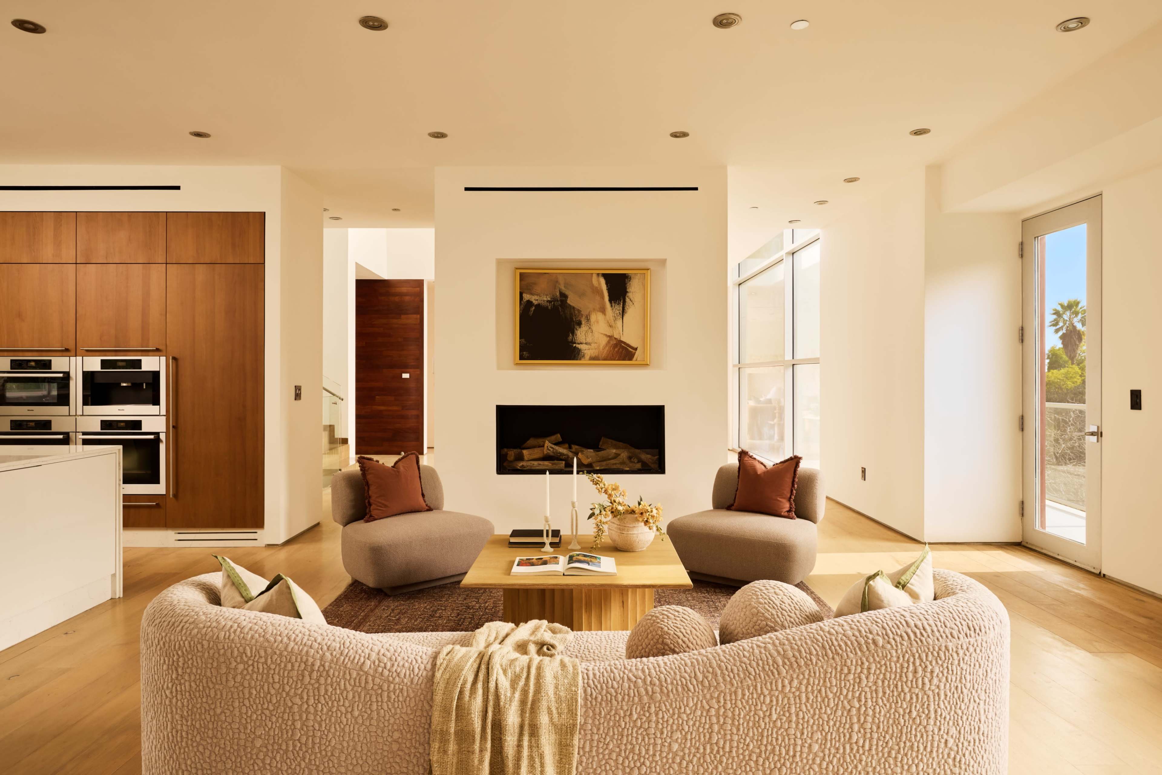 A modern living room features a light-colored, curved sofa, two armchairs, a wooden coffee table, and a large flat-screen television mounted on the wall, with large windows allowing natural light to fill the space.