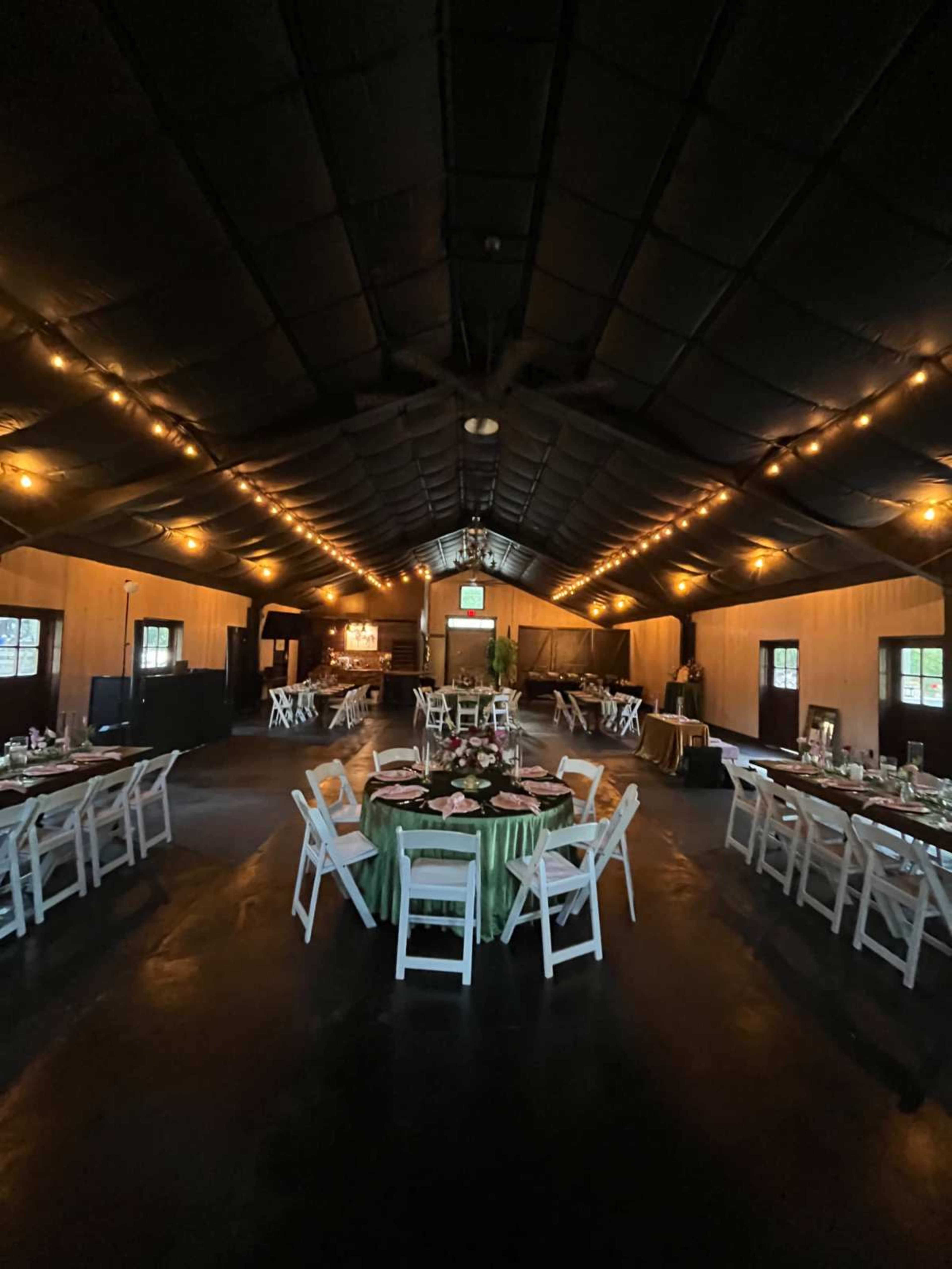 The interior of a large hall features round tables set with white chairs and decorative centerpieces, illuminated by string lights along the ceiling.