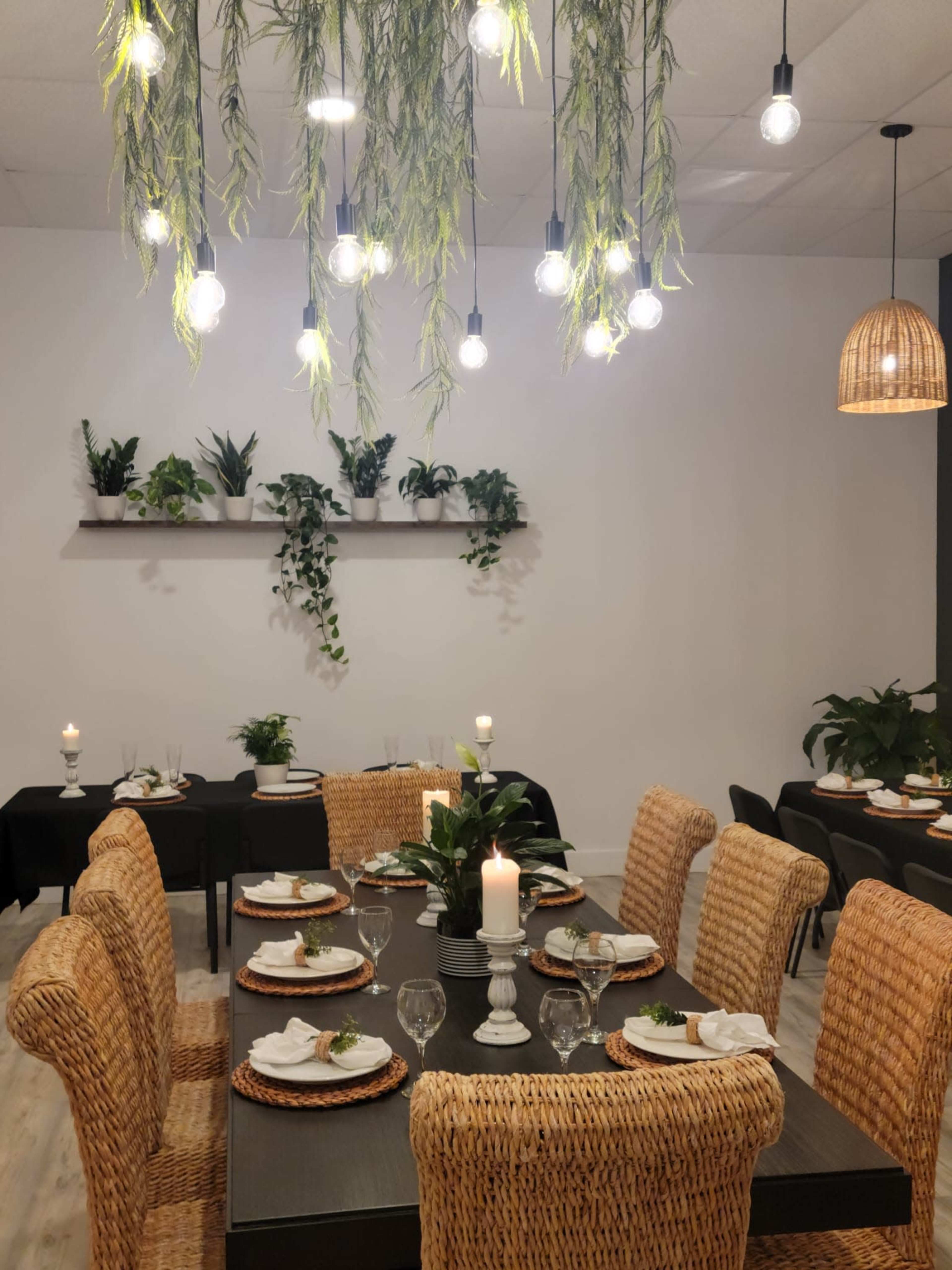 A dining area features a large table set with plates and glasses, surrounded by wicker chairs, with hanging lights and plants adorning the space.