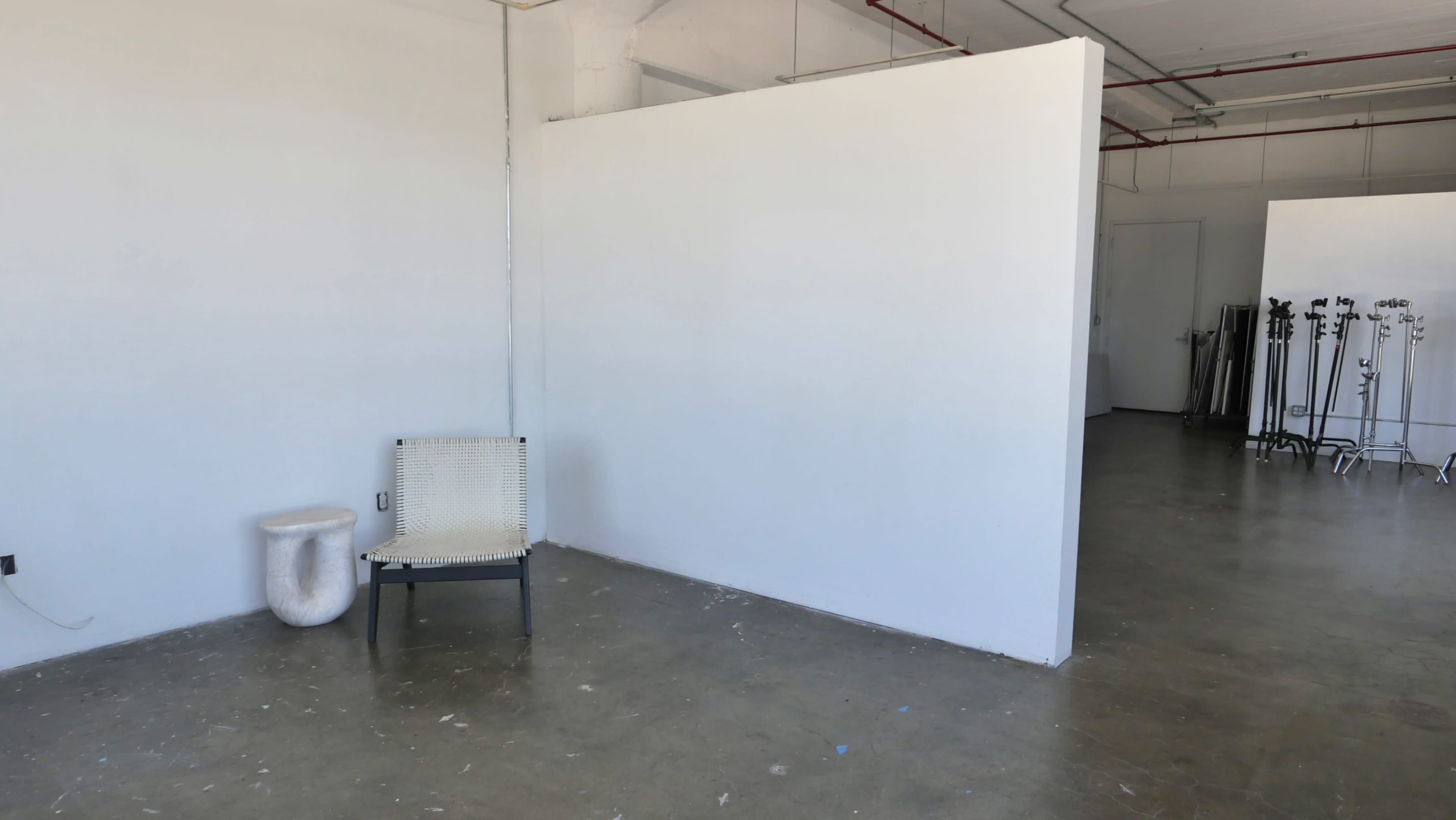 The image shows a spacious, empty room with a gray concrete floor, a white wall partition, a simple chair, and a small stool.