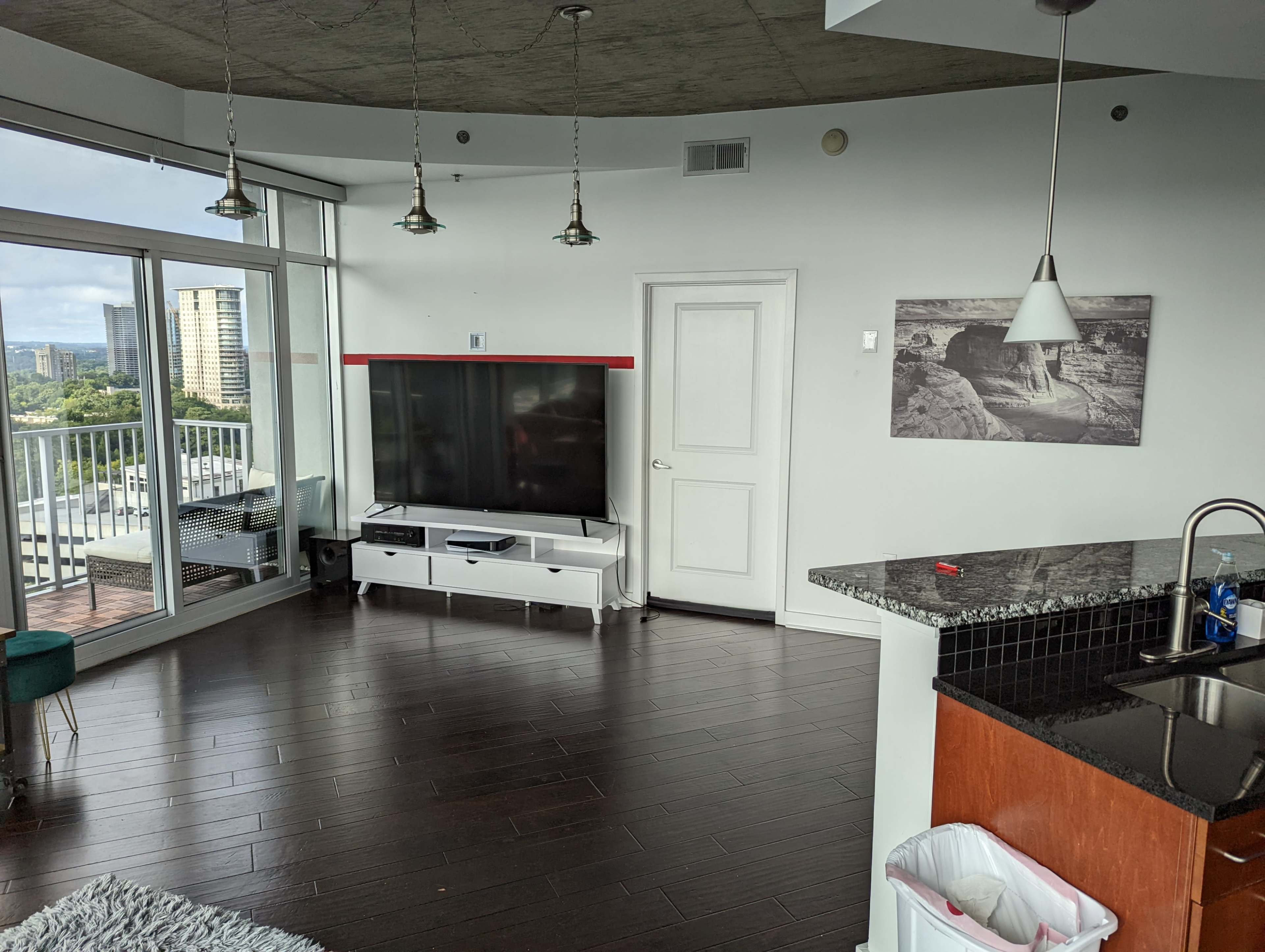 A modern living room featuring a large television mounted on the wall, with a sectional white entertainment unit and a kitchen area in the corner.