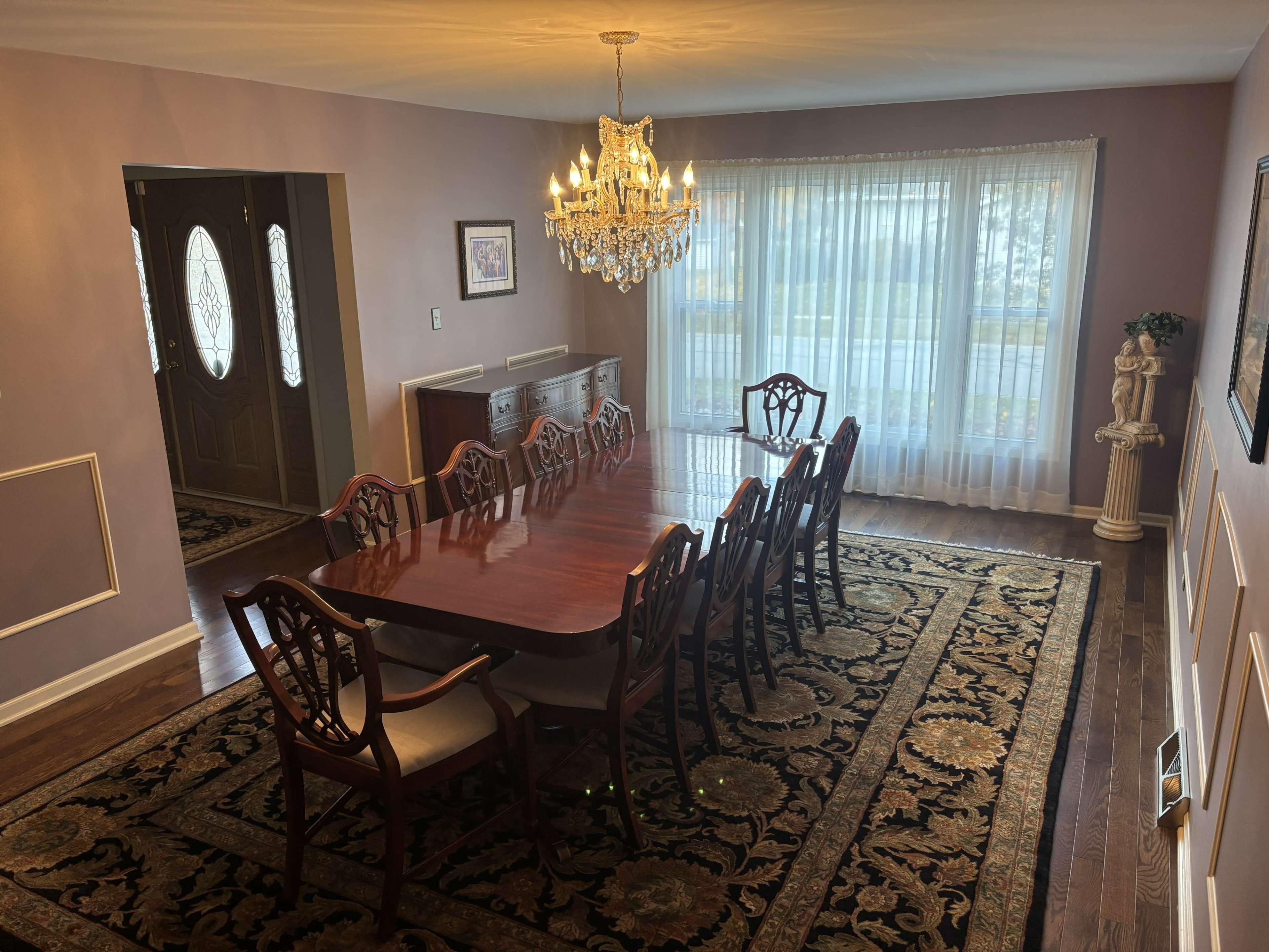 A large dining room features a polished wooden table surrounded by eight chairs, with a chandelier hanging above and large windows allowing natural light to illuminate the space.