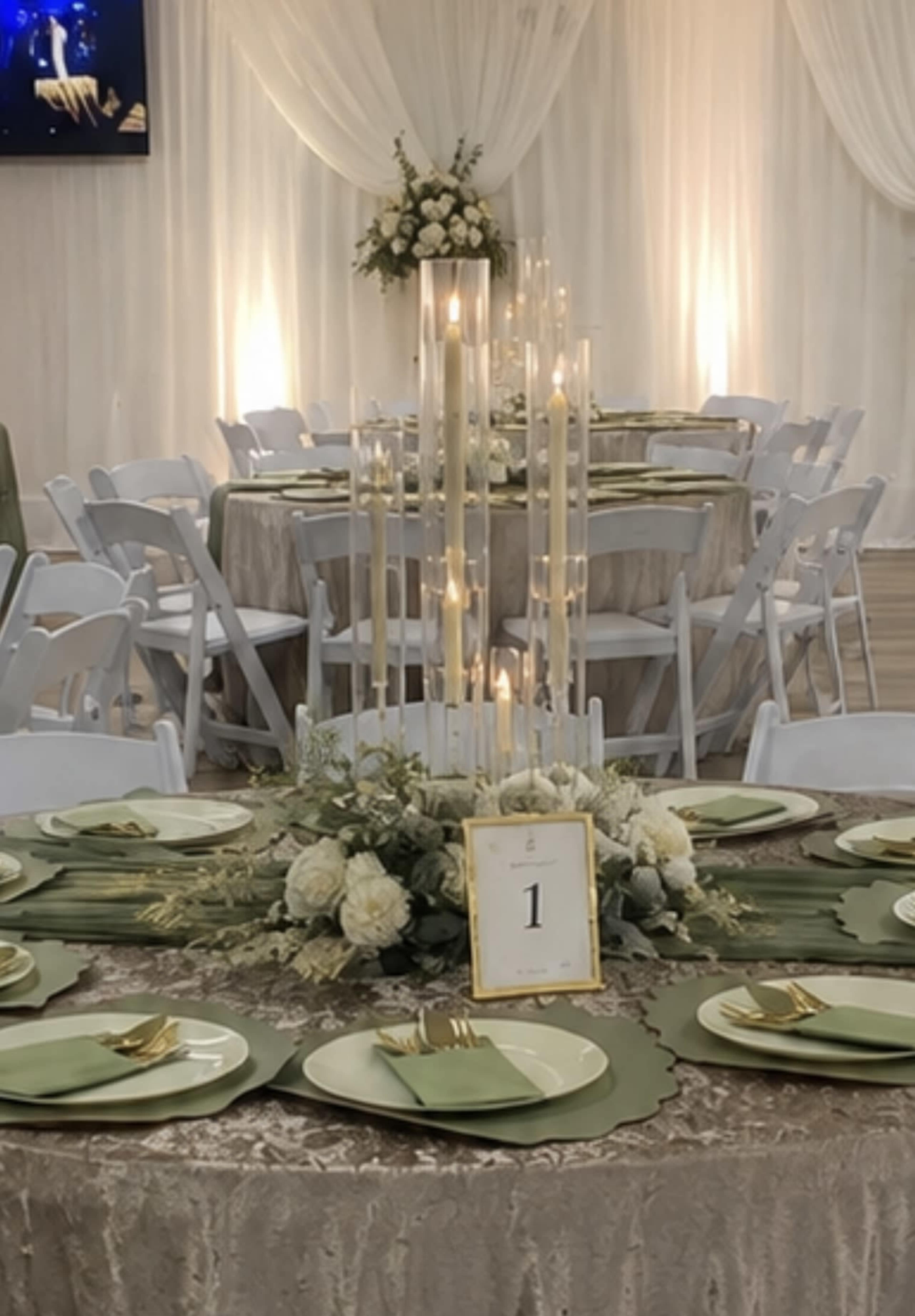 A table is elegantly set with green plates and gold cutlery, featuring a tall candle centerpiece surrounded by flowers.