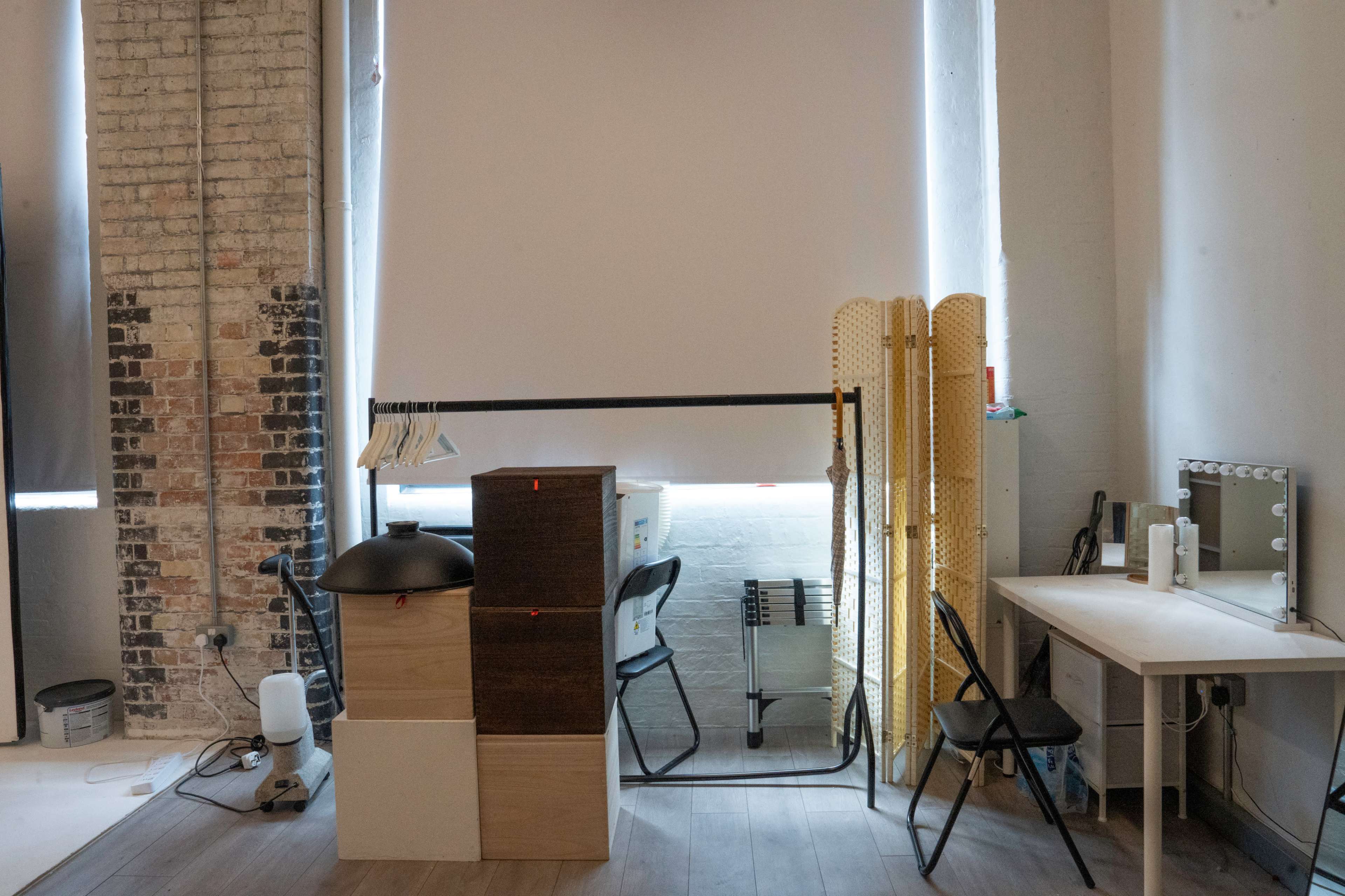 The image shows a minimalist interior with a dressing table, a clothing rack, and various storage boxes against a brick wall.