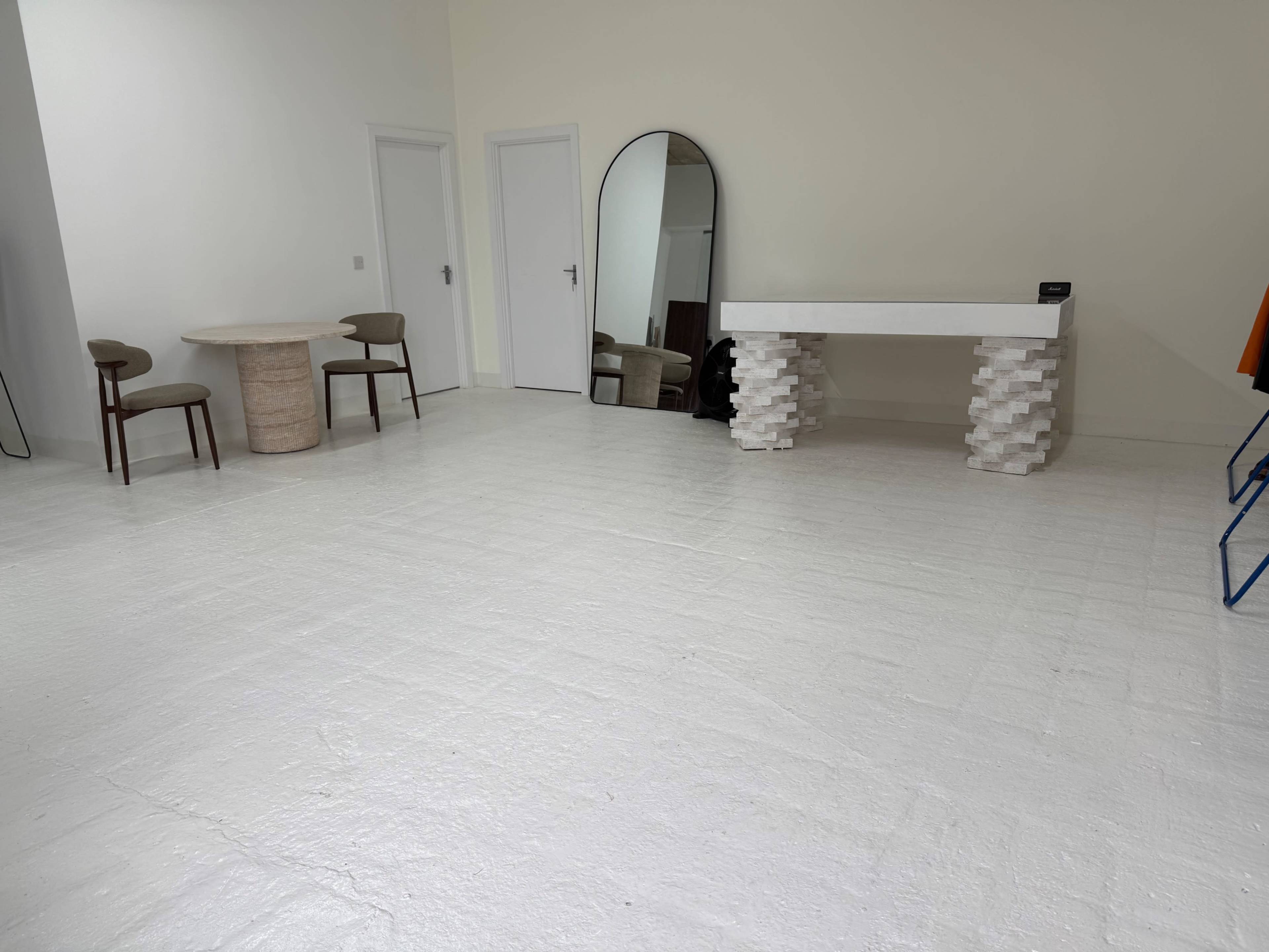 The image shows a minimalist room featuring a round table with two chairs, a console table made of blocks, an oval mirror, and a large empty floor space.