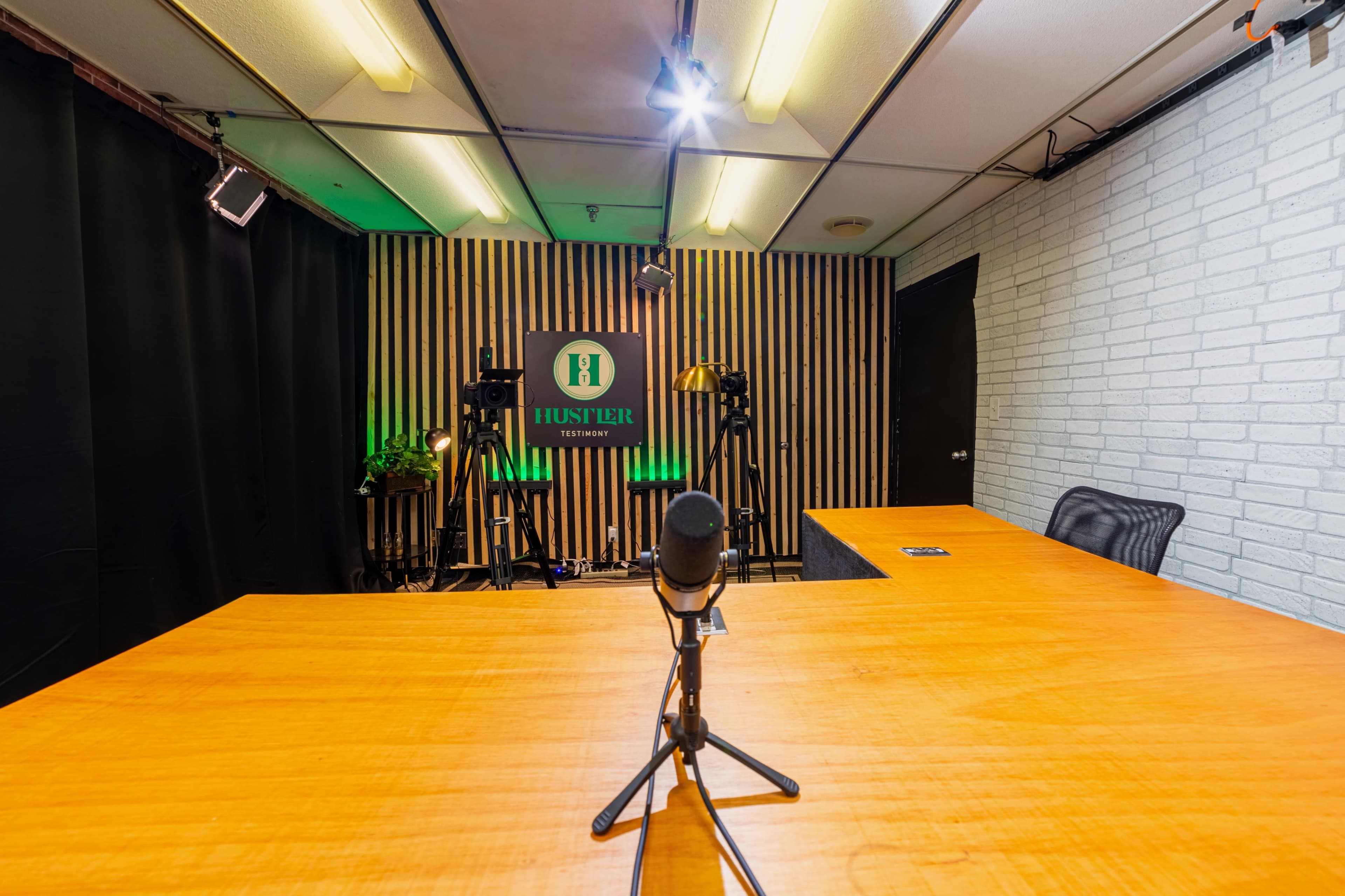 The image shows a recording studio with a large wooden table, a microphone on a stand, and various filming equipment arranged in the background.