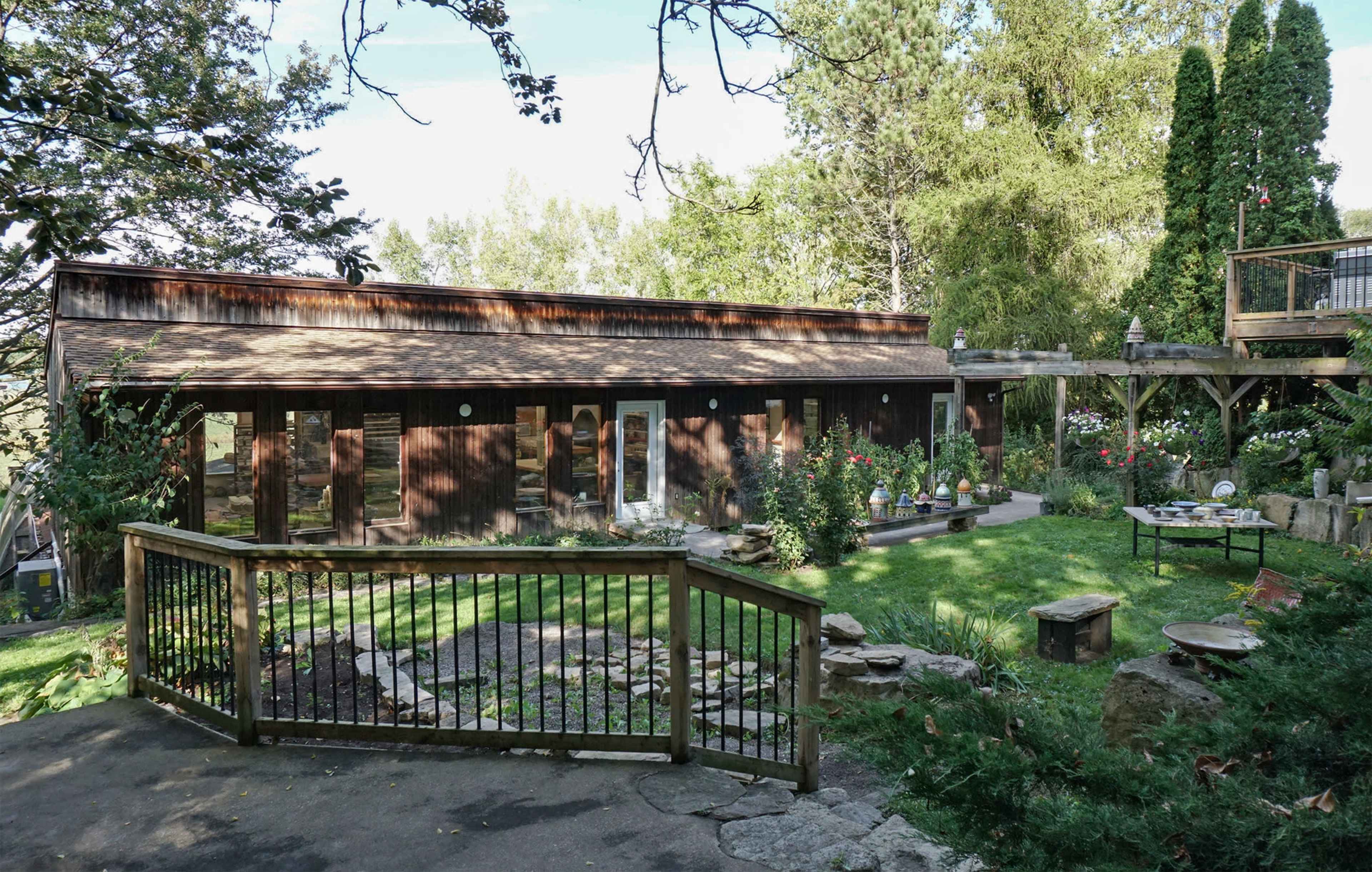 The image shows a wooden house surrounded by greenery and a well-maintained garden with various plants and outdoor seating areas.