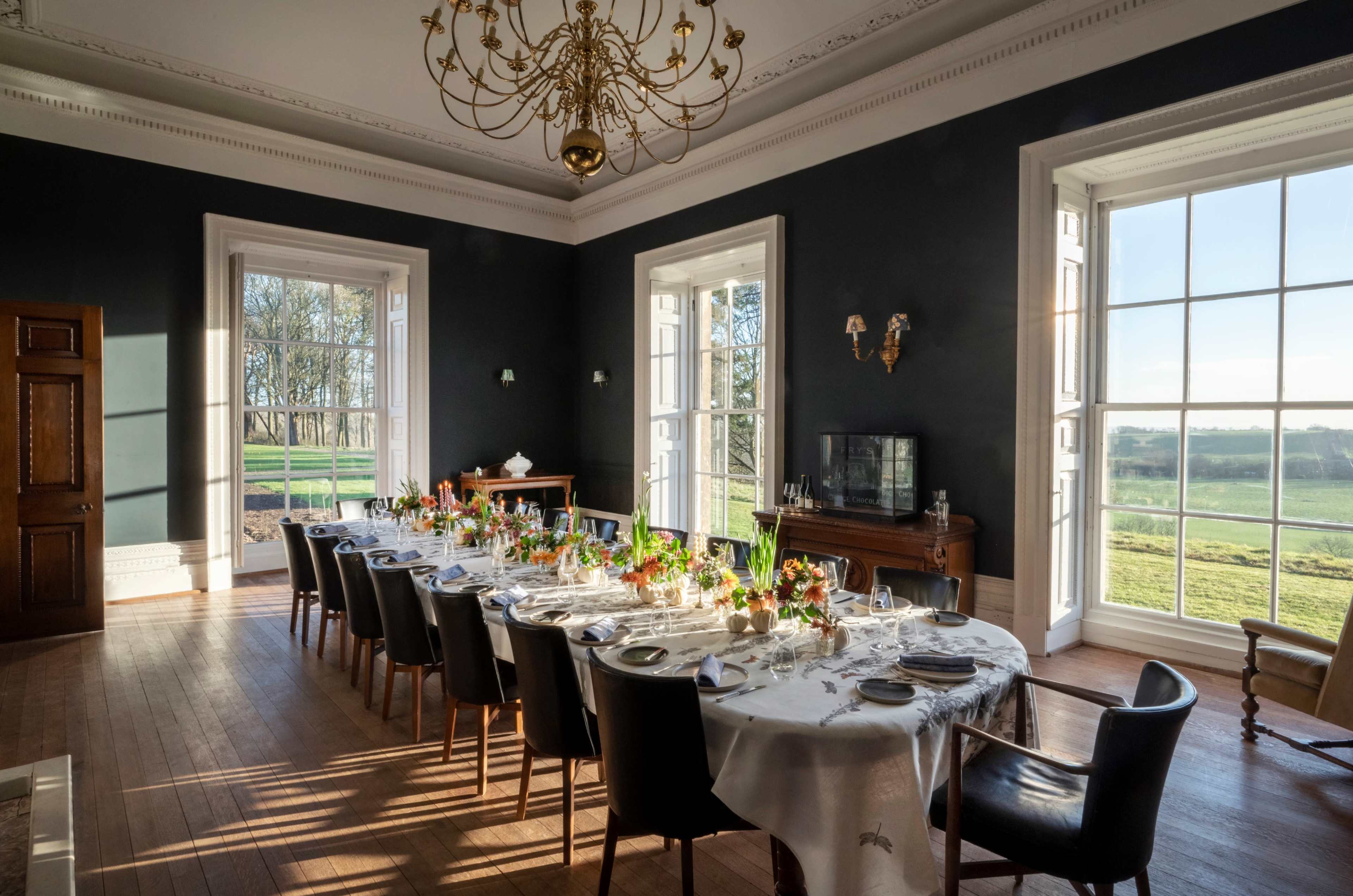 A long dining table adorned with floral centerpieces is set against a backdrop of large windows that let in natural light, revealing a green landscape outside.