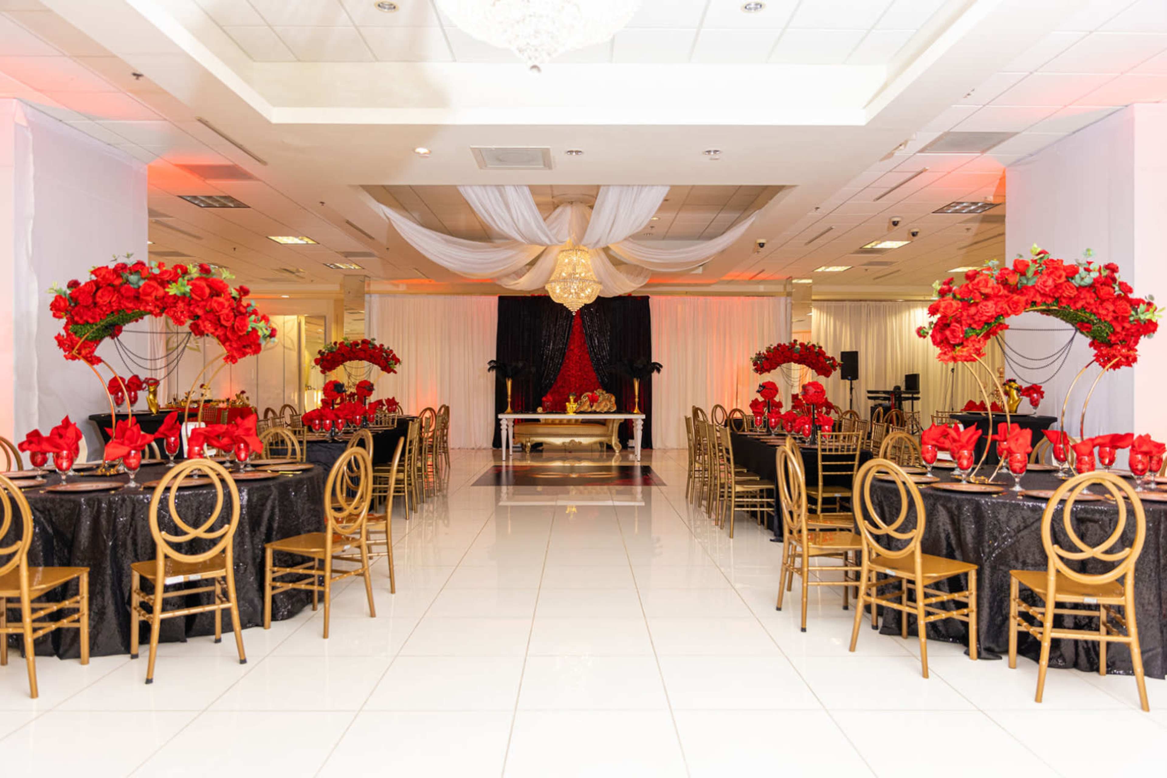 The image shows a banquet hall elegantly decorated with red floral arrangements and black tablecloths, featuring round tables with golden chairs.