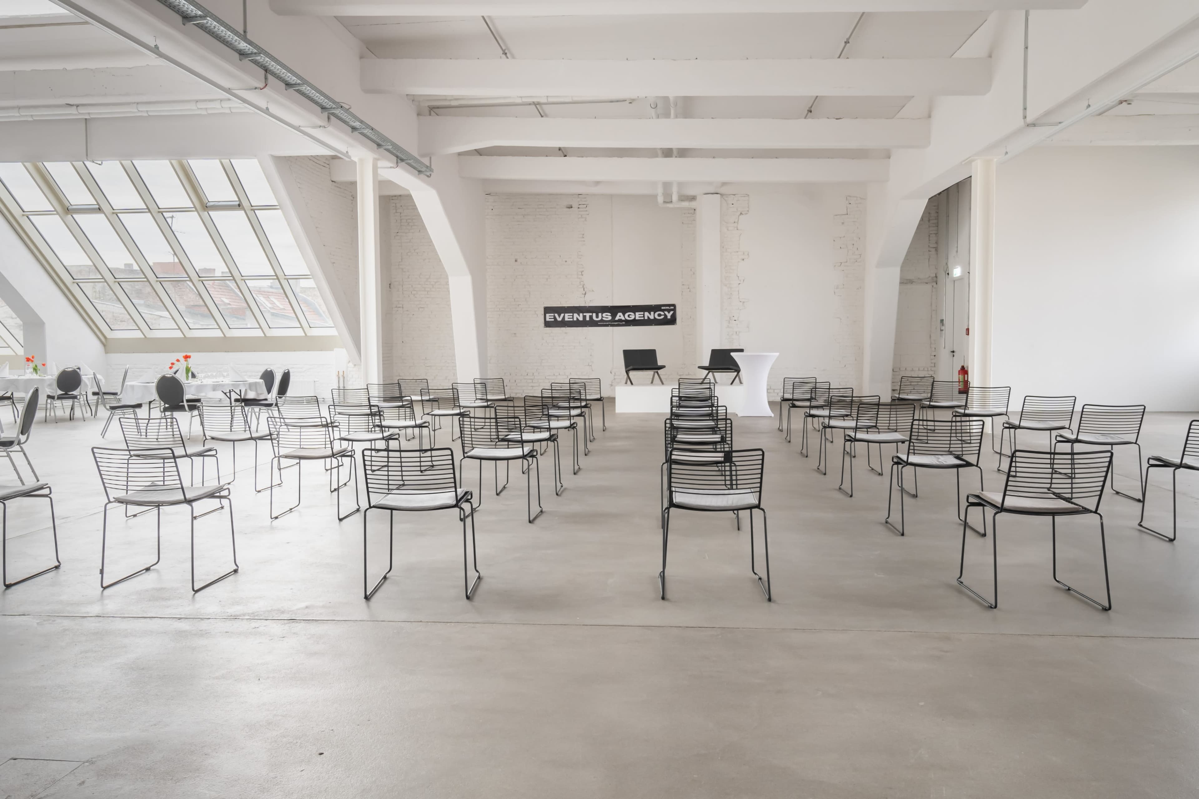 A spacious, minimalist conference room with rows of empty black wire chairs arranged for an event, featuring a backdrop that reads "EVENTUS AGENCY."