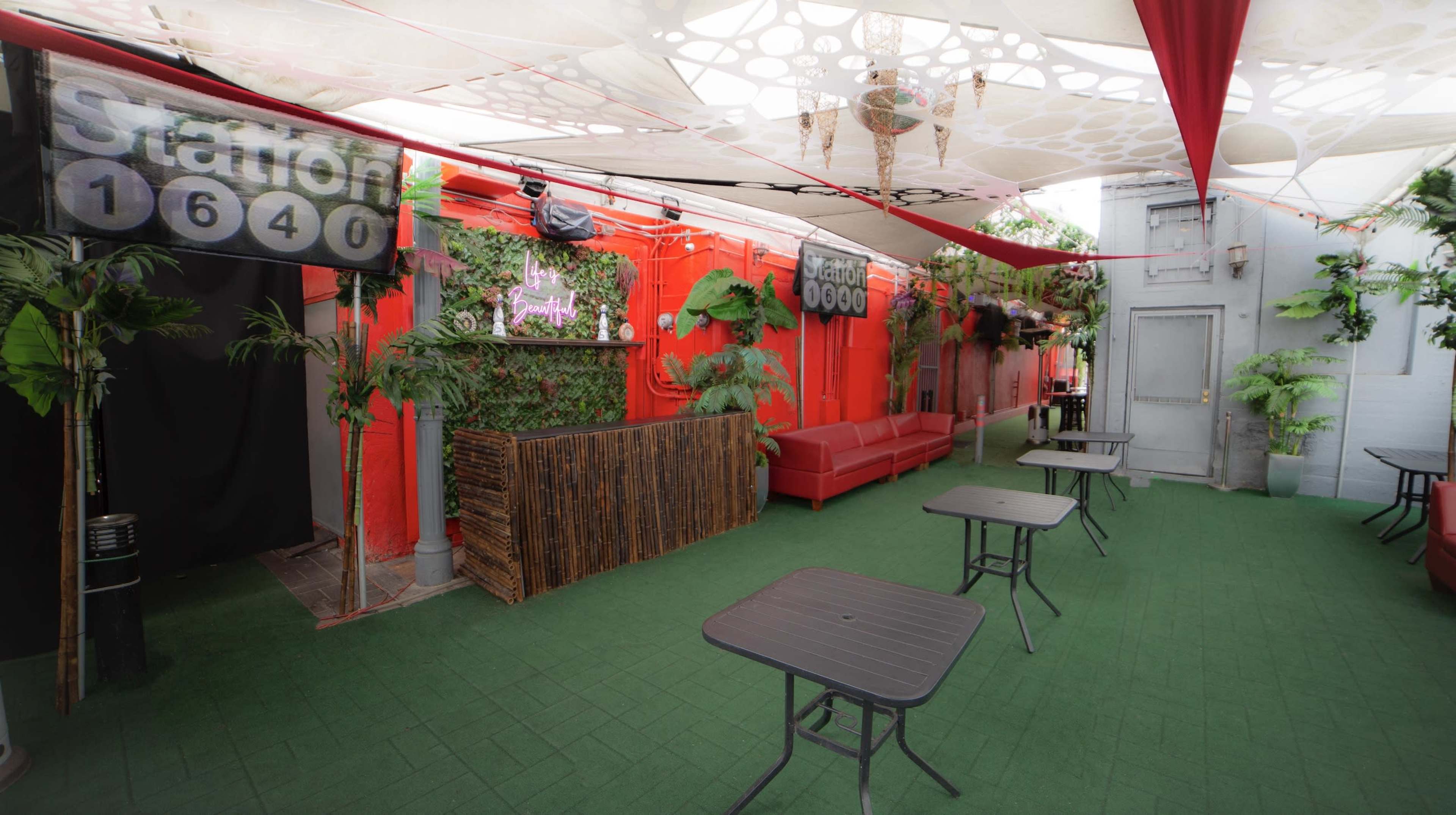 A vibrant outdoor space featuring red walls, green foliage, a seating area with a red couch, and several tables arranged on a green floor.