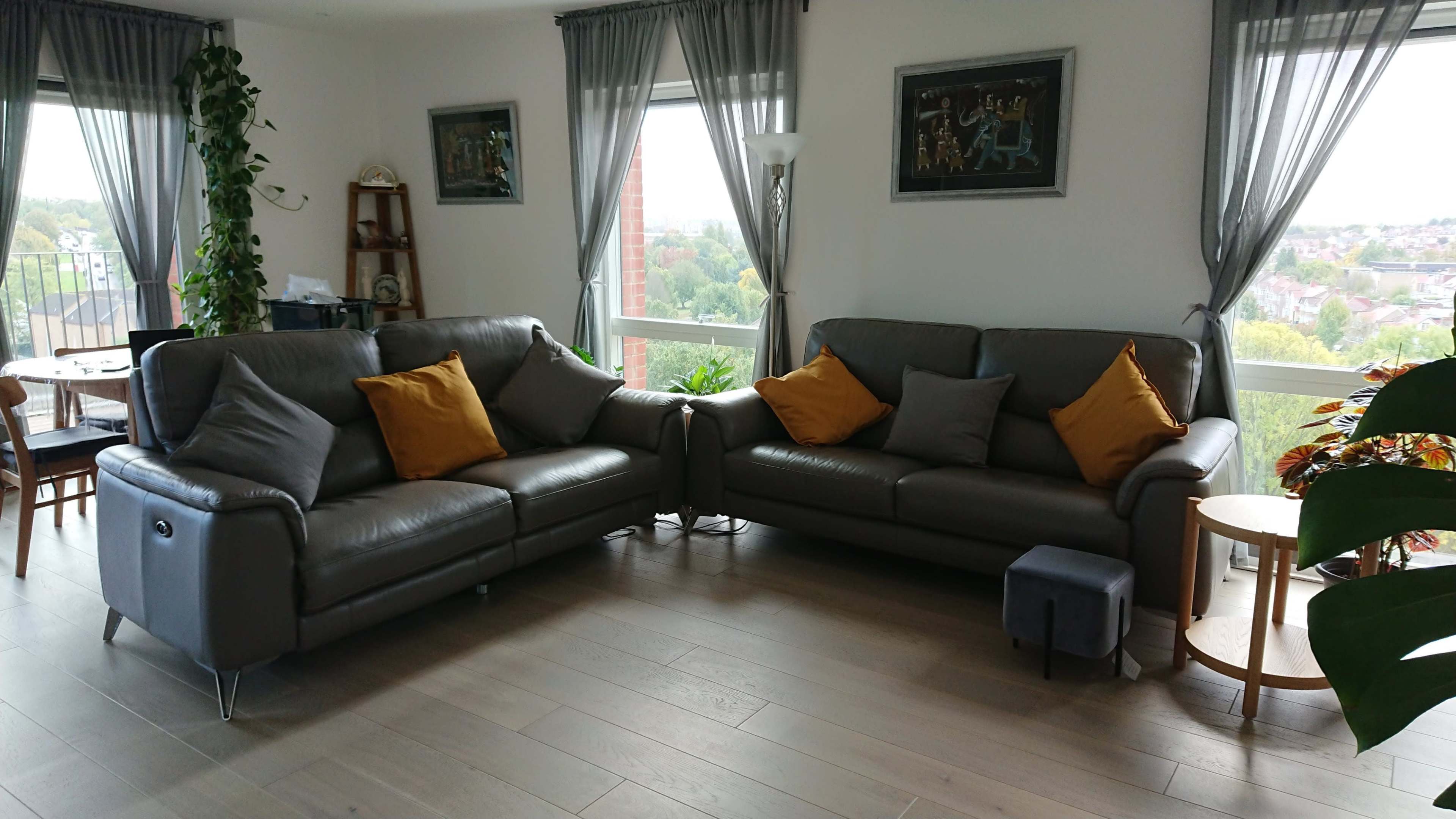 A living room features two gray leather sofas with orange and gray cushions arranged around a small table, with large windows providing a view of the outdoors.