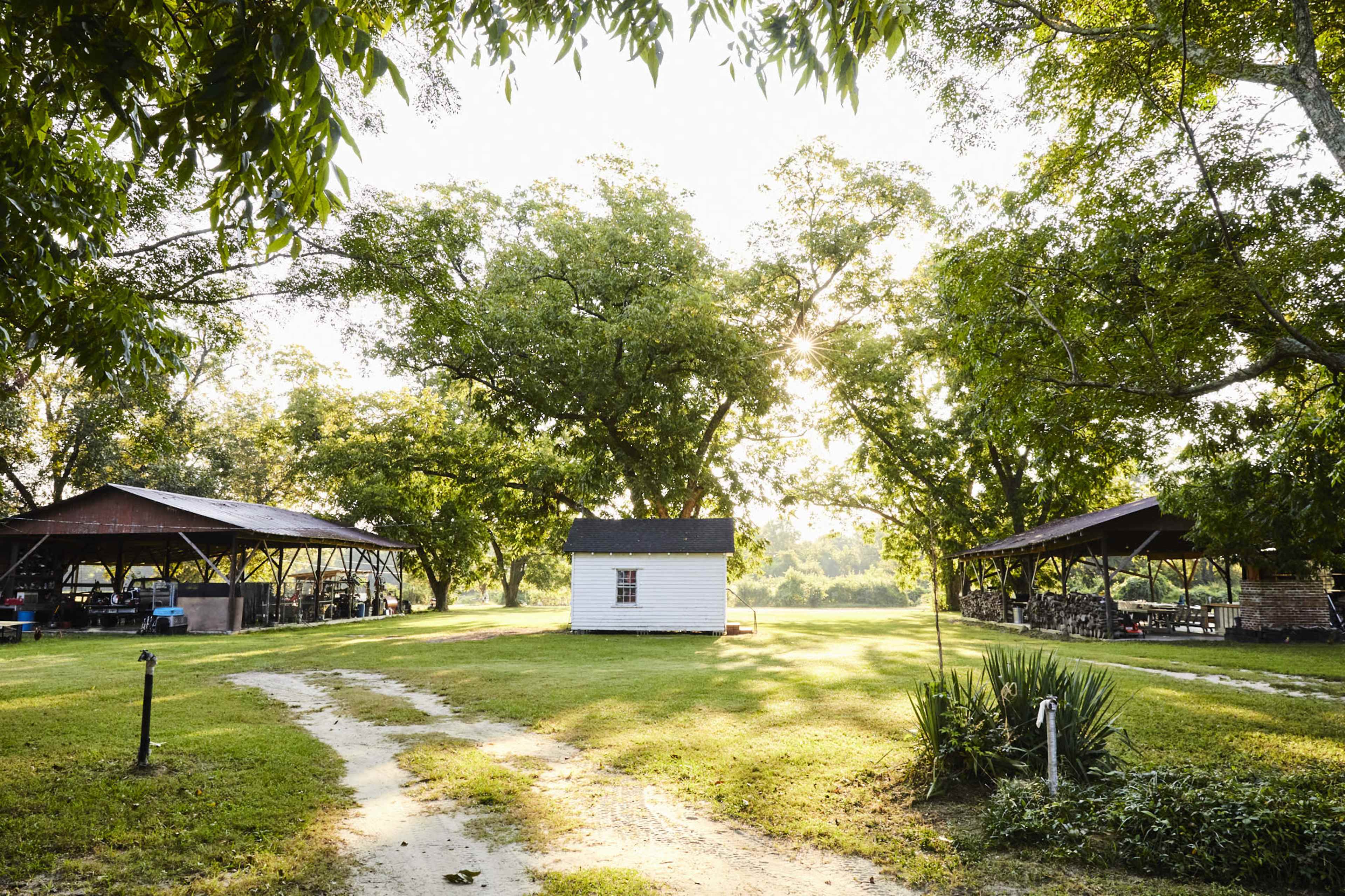 A small white shed is situated on a grassy area surrounded by large trees and two covered structures in a rural setting.