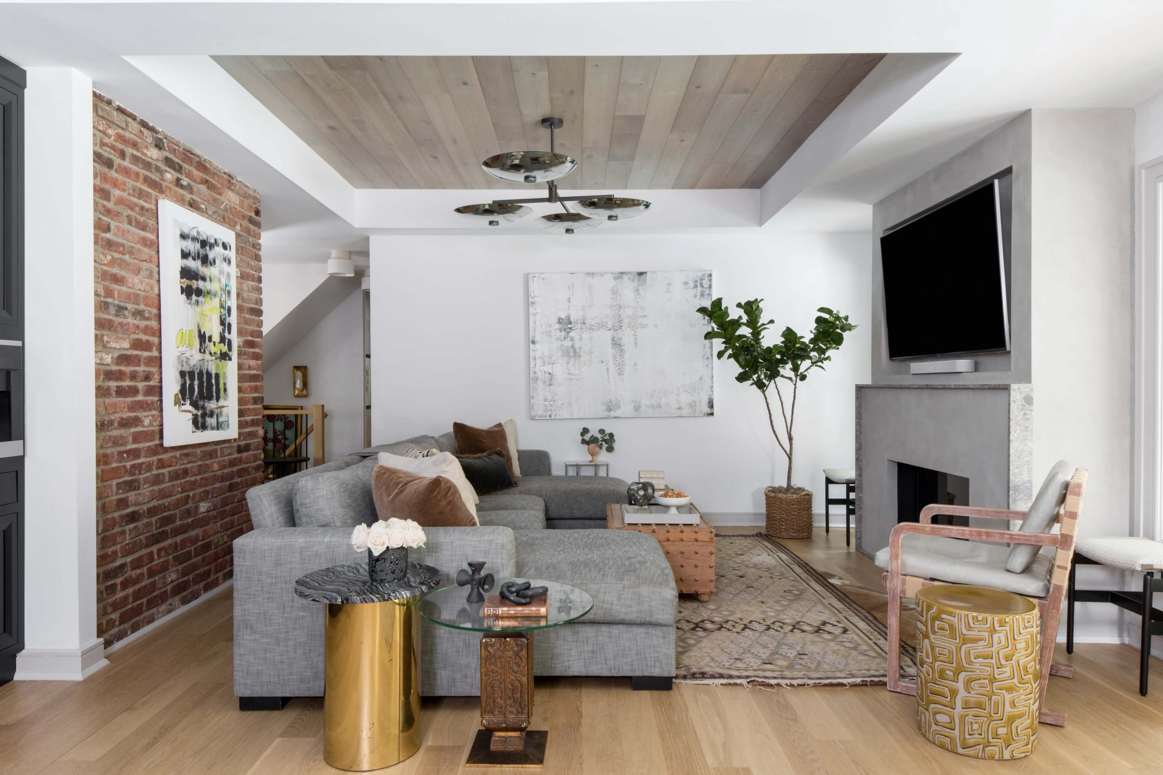 A modern living room featuring a gray sectional sofa, a coffee table, a wall-mounted TV, and a lush green plant, situated beside exposed brick and wooden ceiling elements.
