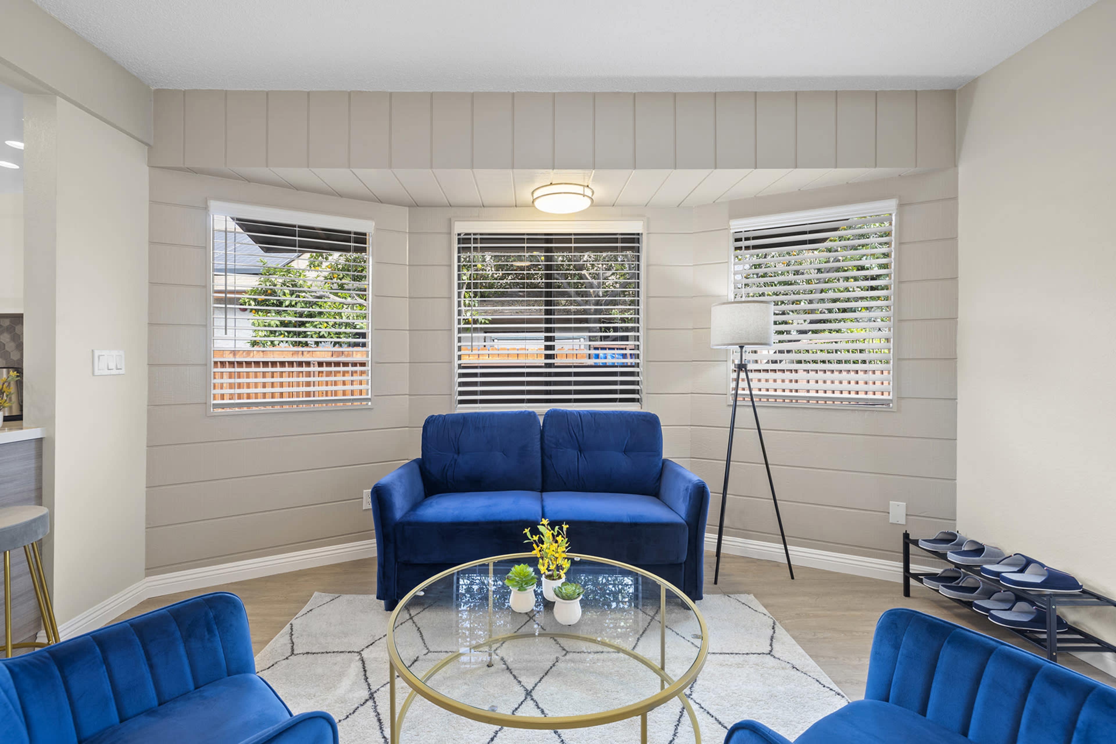 A cozy living room with a blue couch and armchairs arranged around a circular glass coffee table, surrounded by windows with blinds.