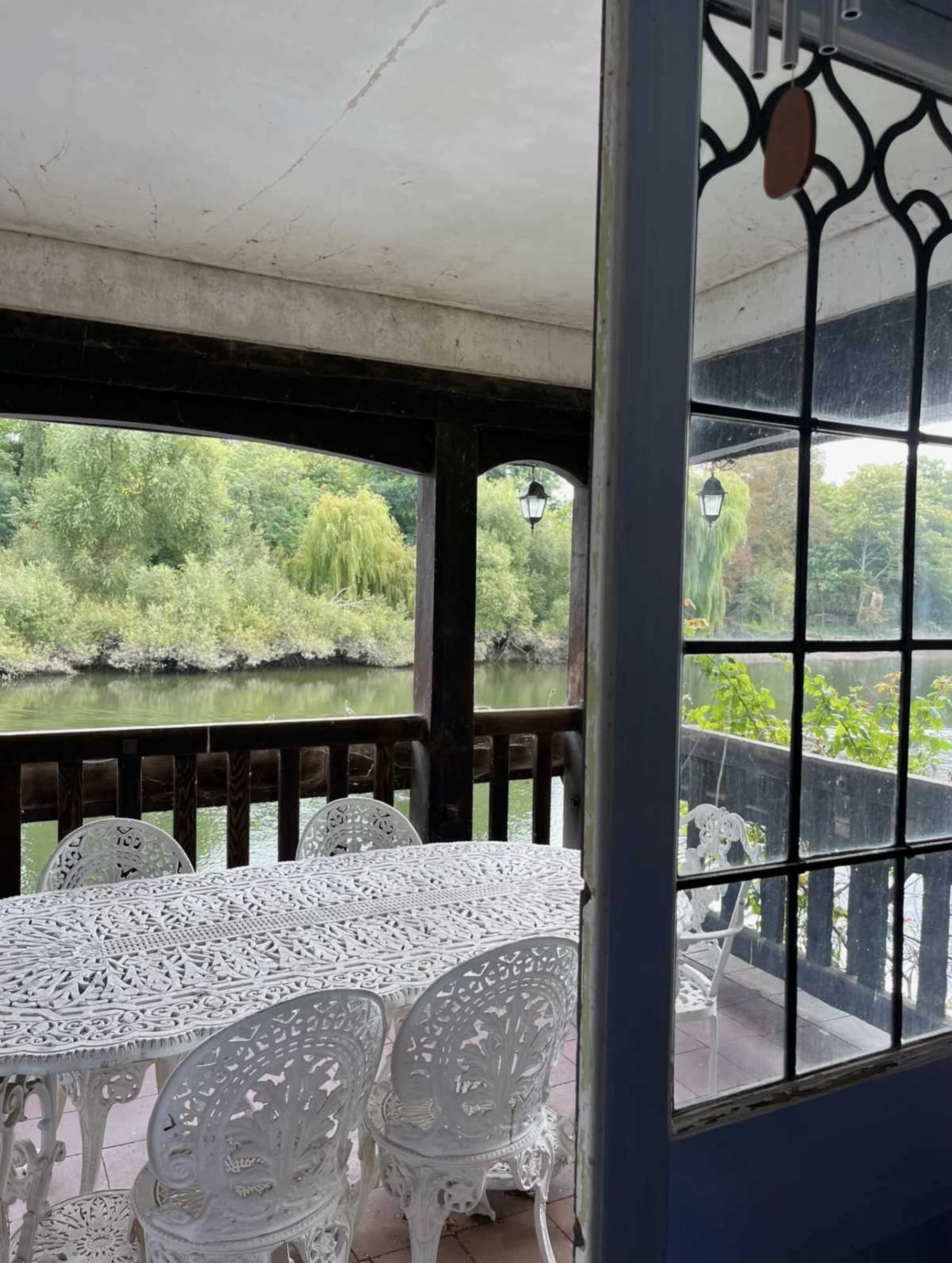 A white wrought iron table and chairs are set on a covered porch overlooking a calm river surrounded by greenery.