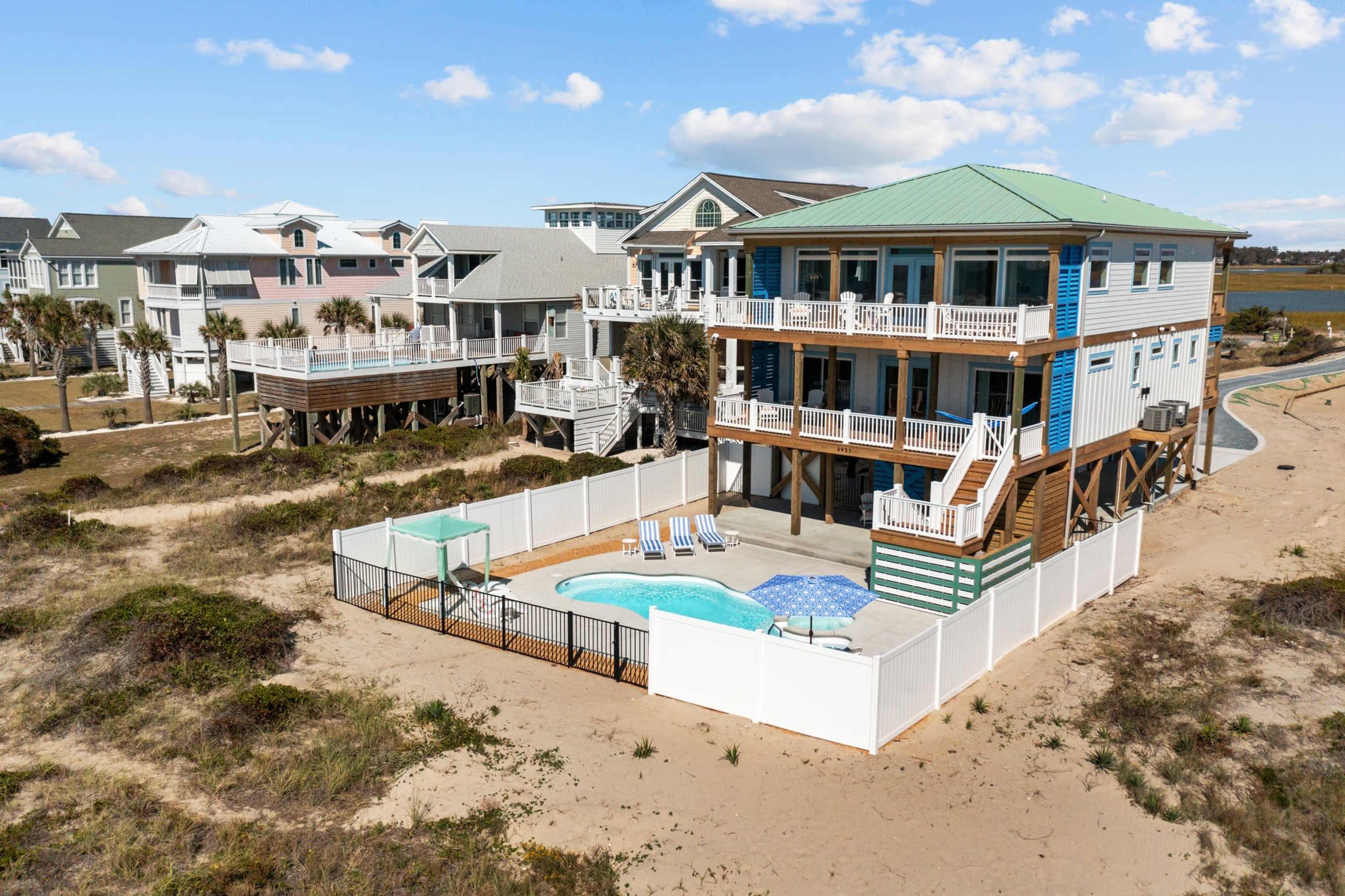A large beach house with a green roof and multiple balconies stands adjacent to a fenced pool area on a sandy lot.