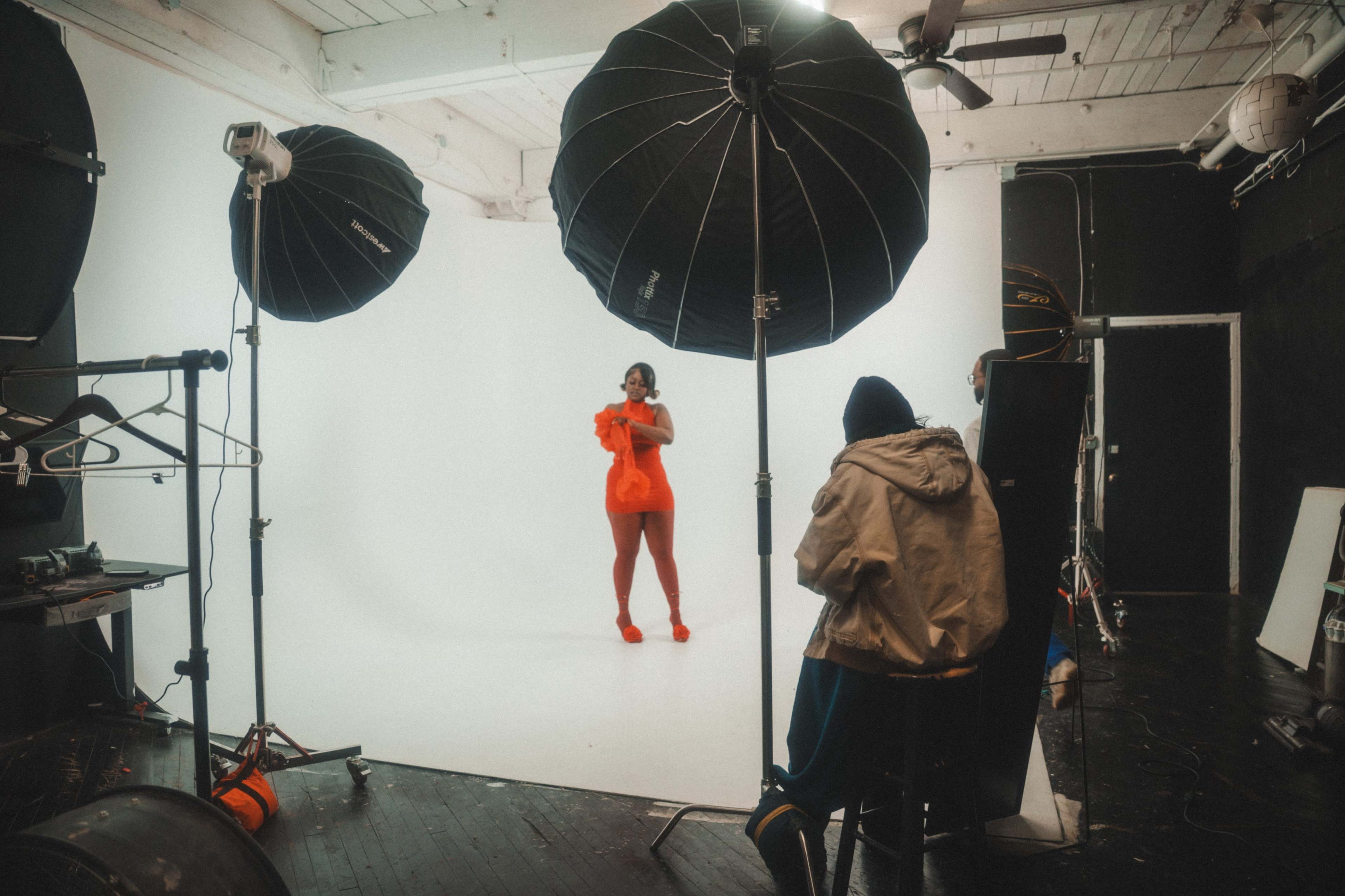 A model in a bright outfit poses on a white backdrop in a photography studio, surrounded by large softboxes and a seated person observing the shoot.