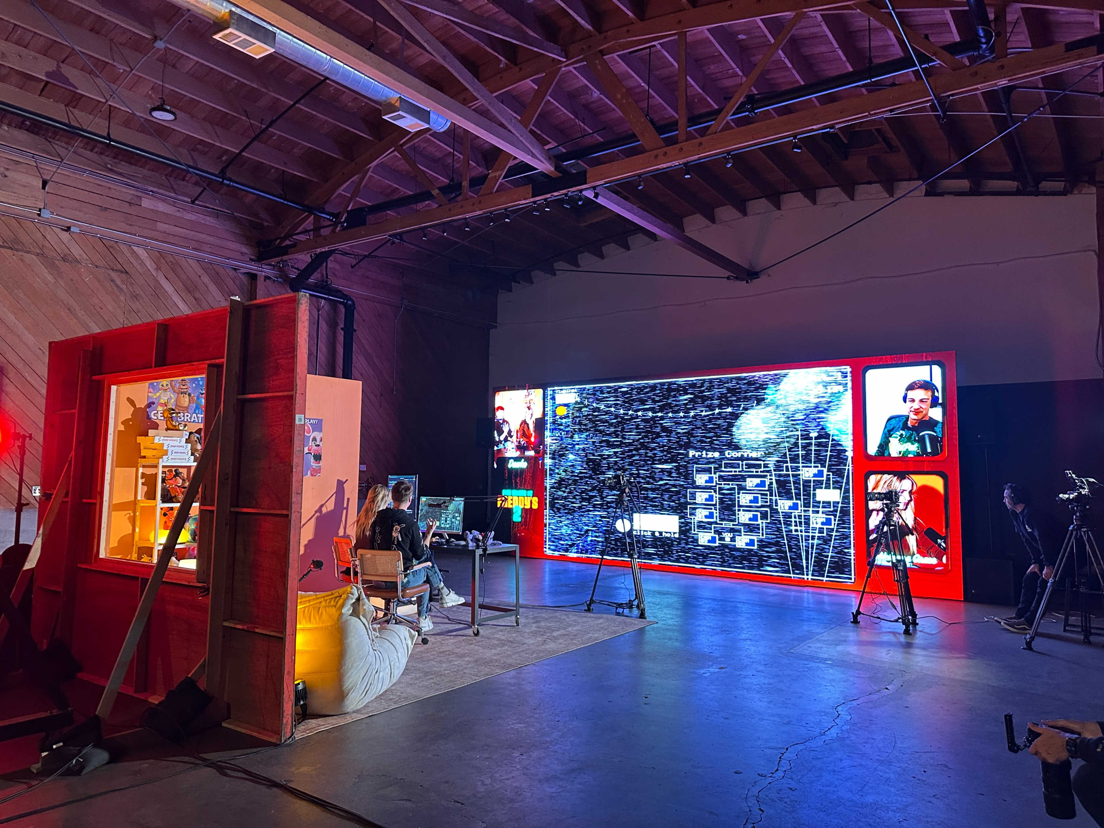 A person sits at a computer in a studio setting, with a large screen displaying digital graphics and various interactive elements.