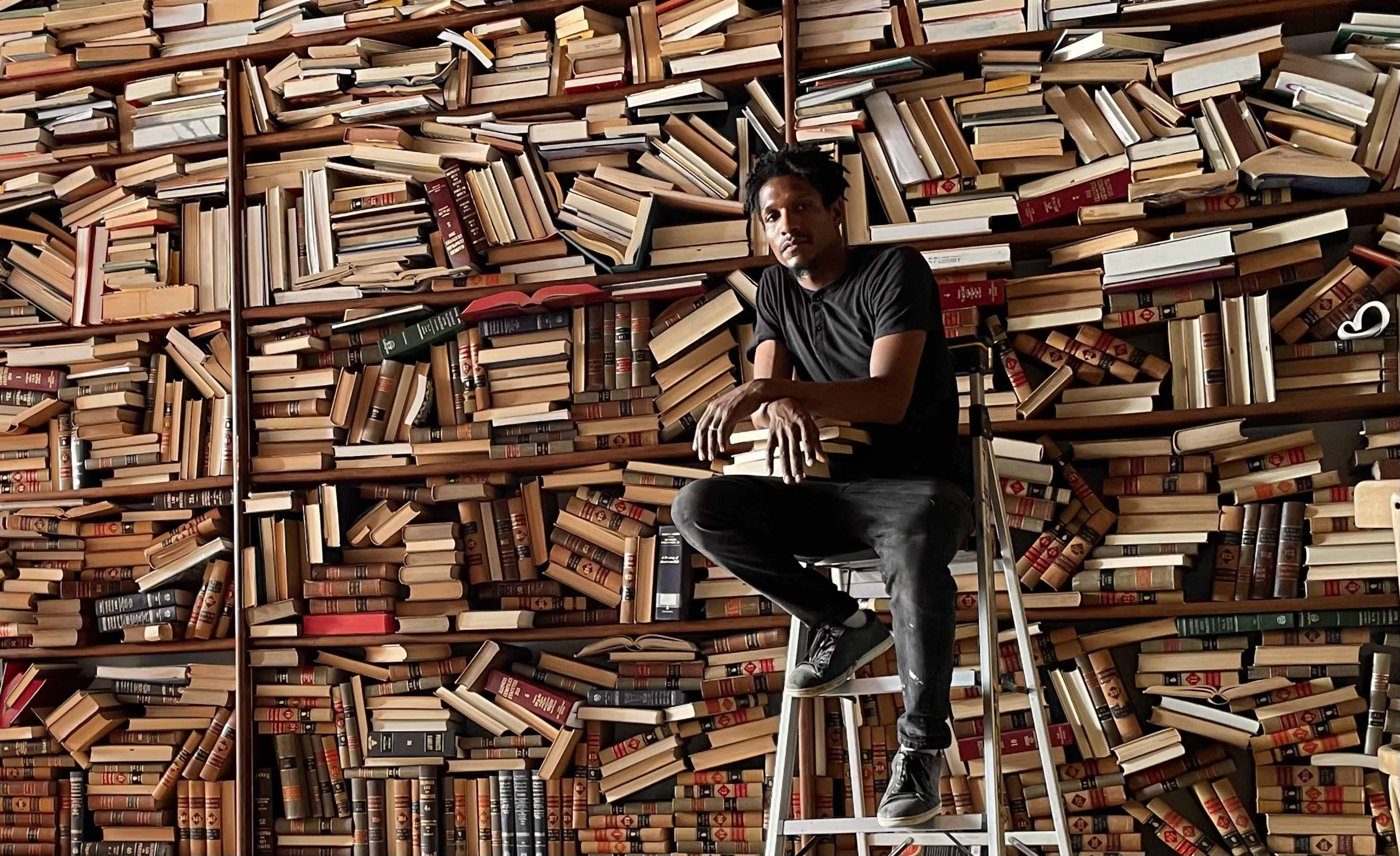 A person sits on a stool in front of a wall densely covered with stacked books.