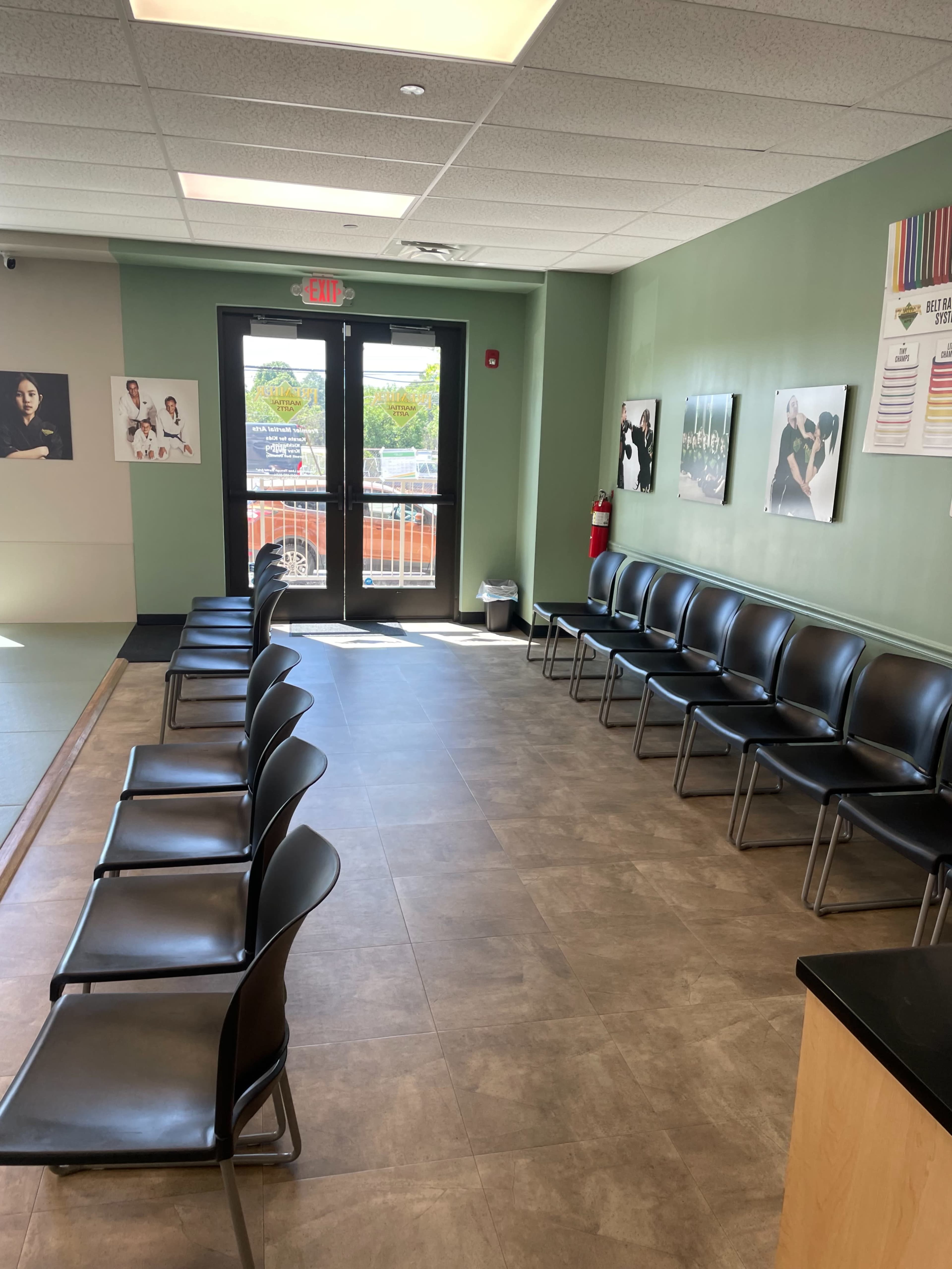 The image shows a waiting area with a row of black chairs along the wall, natural light coming in through a glass door, and several framed posters hanging on the walls.