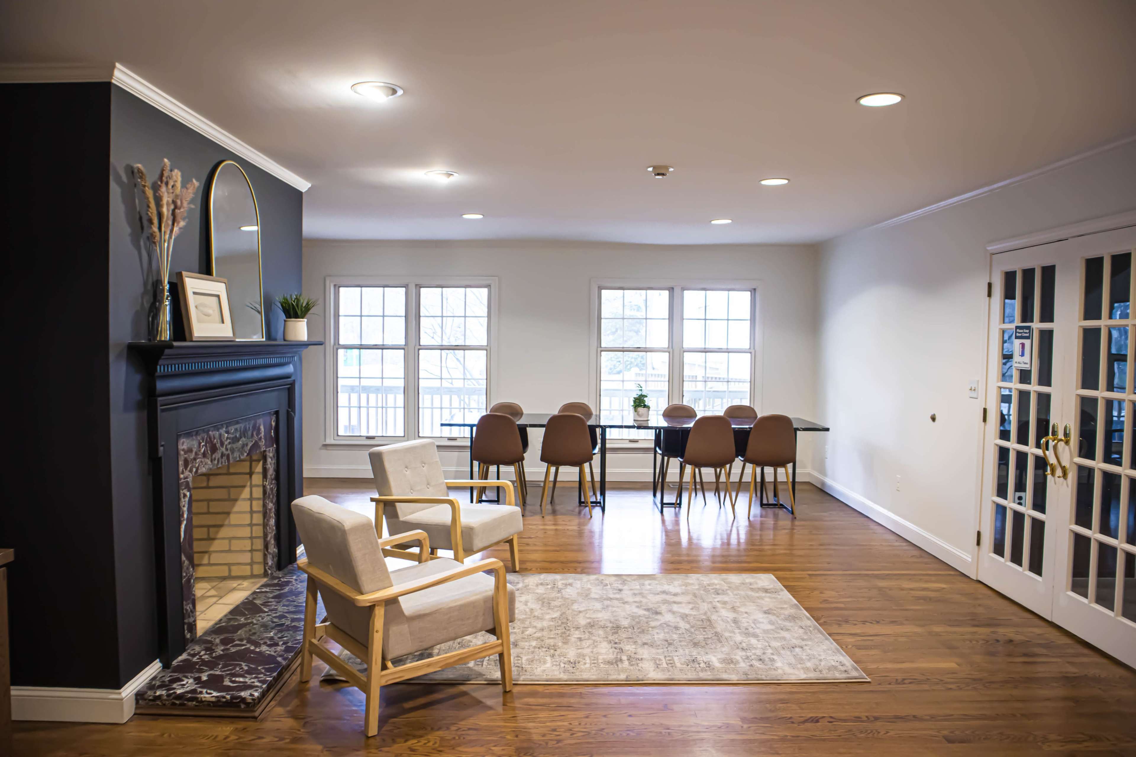 A spacious living area featuring a white chair and a fireplace on one side, with a dining table and chairs set up in front of large windows on the opposite side.