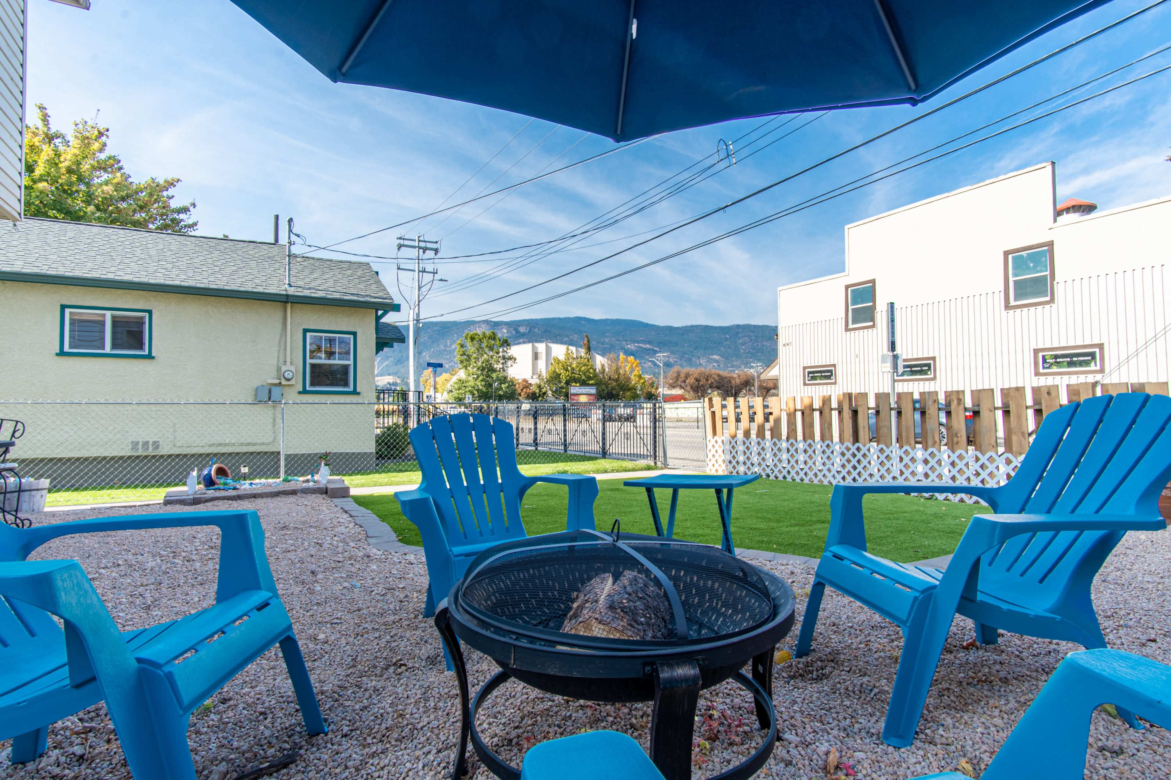 A gravel yard features a fire pit surrounded by blue Adirondack chairs, with a backdrop of mountains and buildings under a clear sky.