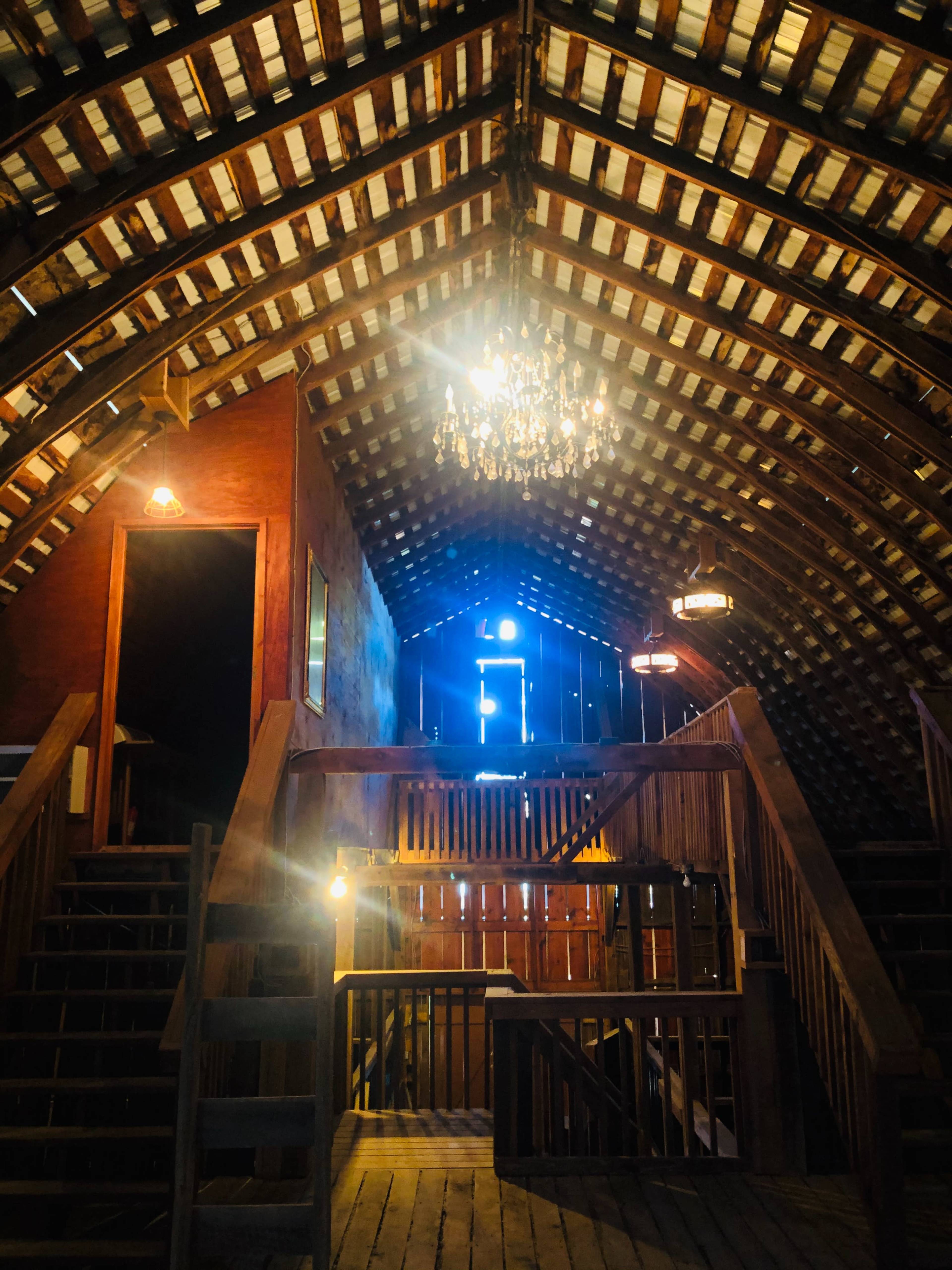 The interior of a wooden barn features a high, beamed ceiling with a chandelier, staircases leading to an upper level, and light streaming through an open door.