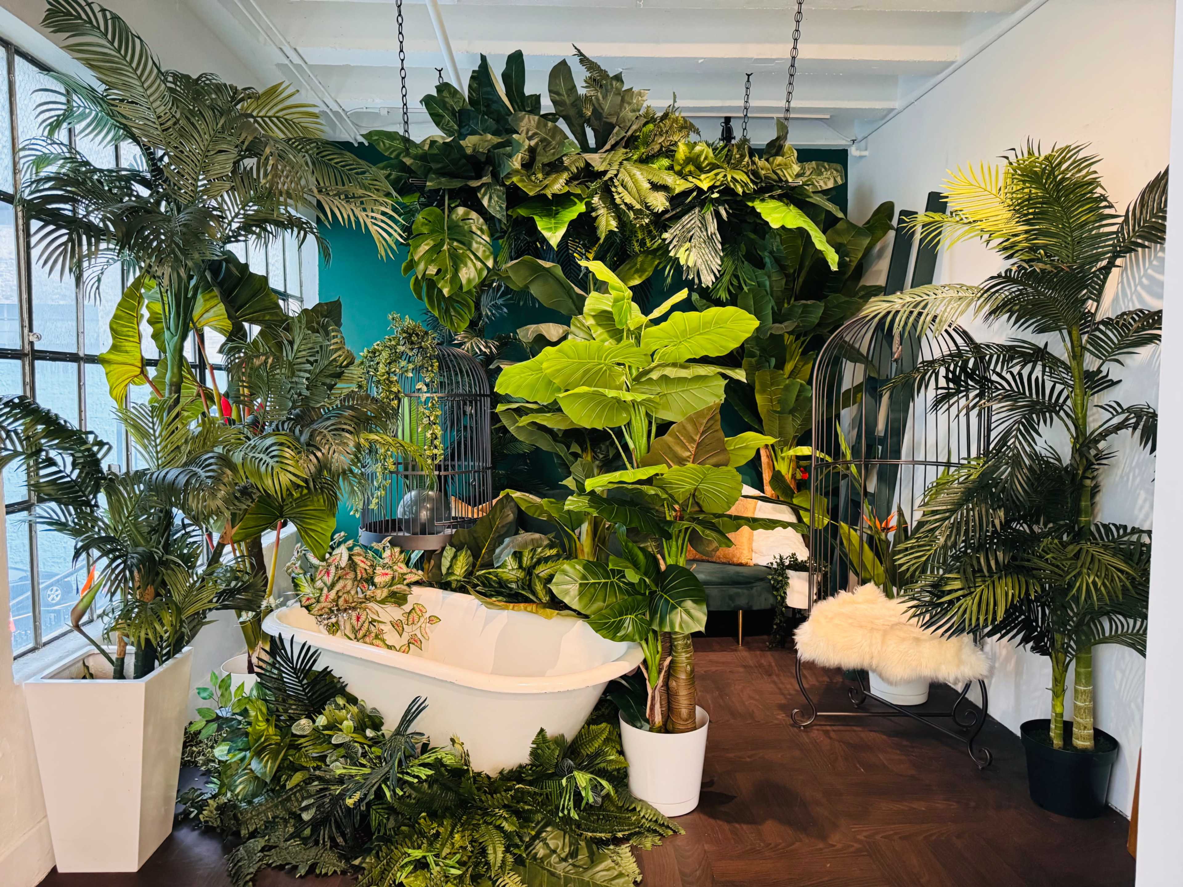 The scene features a lush indoor space filled with various plants, including large leafy tropical varieties, surrounding a freestanding bathtub and a couple of seating areas.