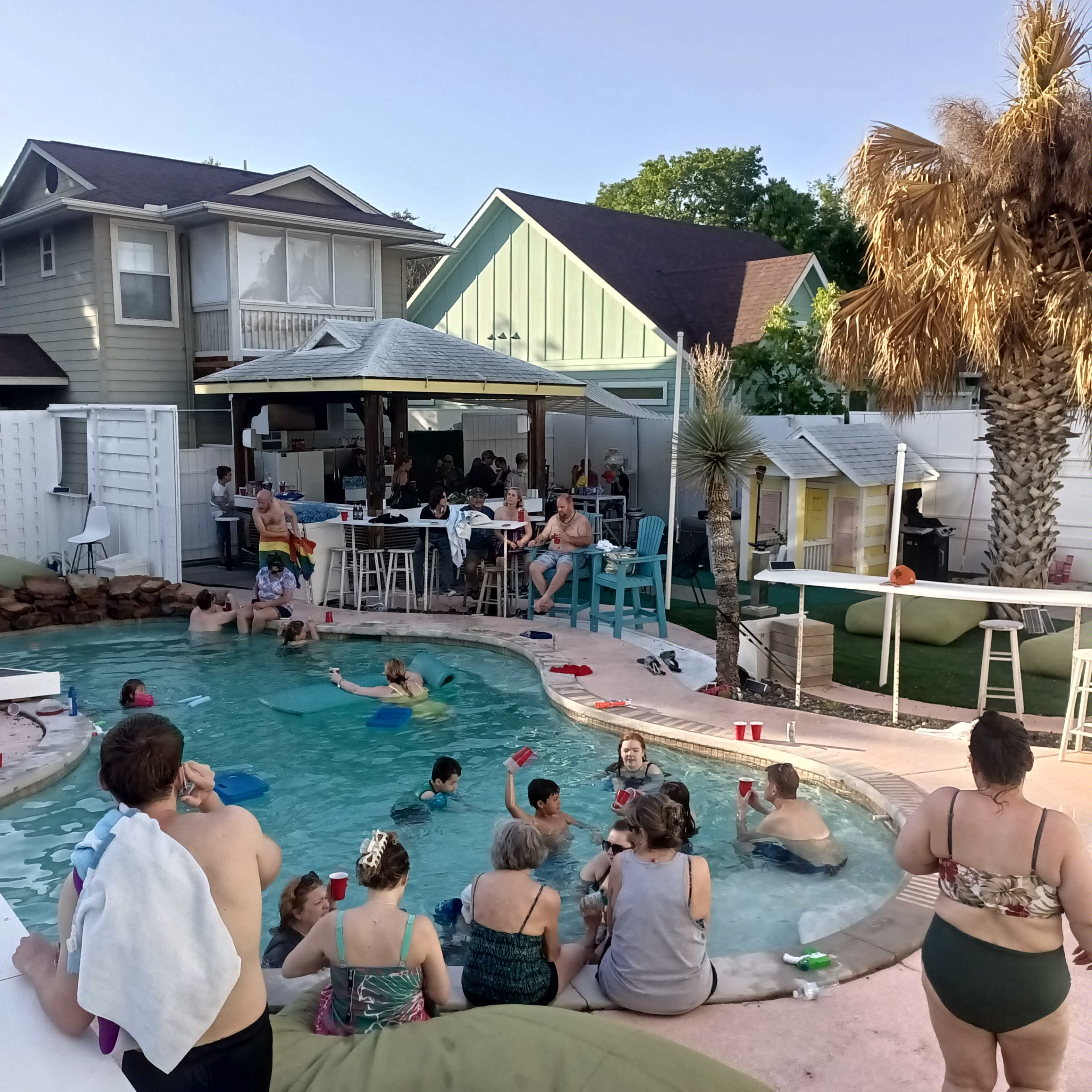 A lively gathering of people is enjoying a pool party in a backyard setting, with some sitting around the pool and others in the water.