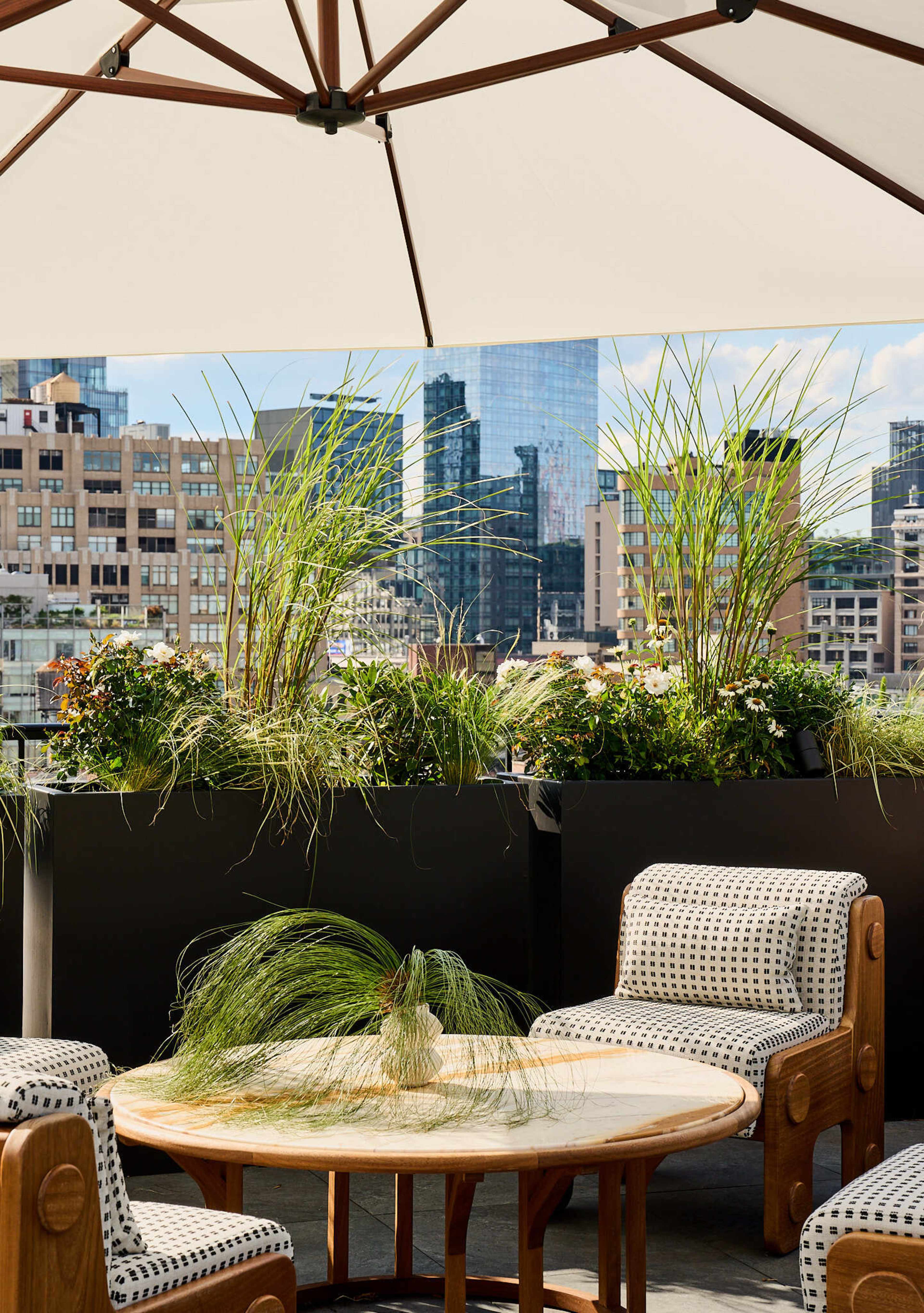 A terrace features potted plants and a round table with two patterned chairs, set against a backdrop of city skyscrapers.