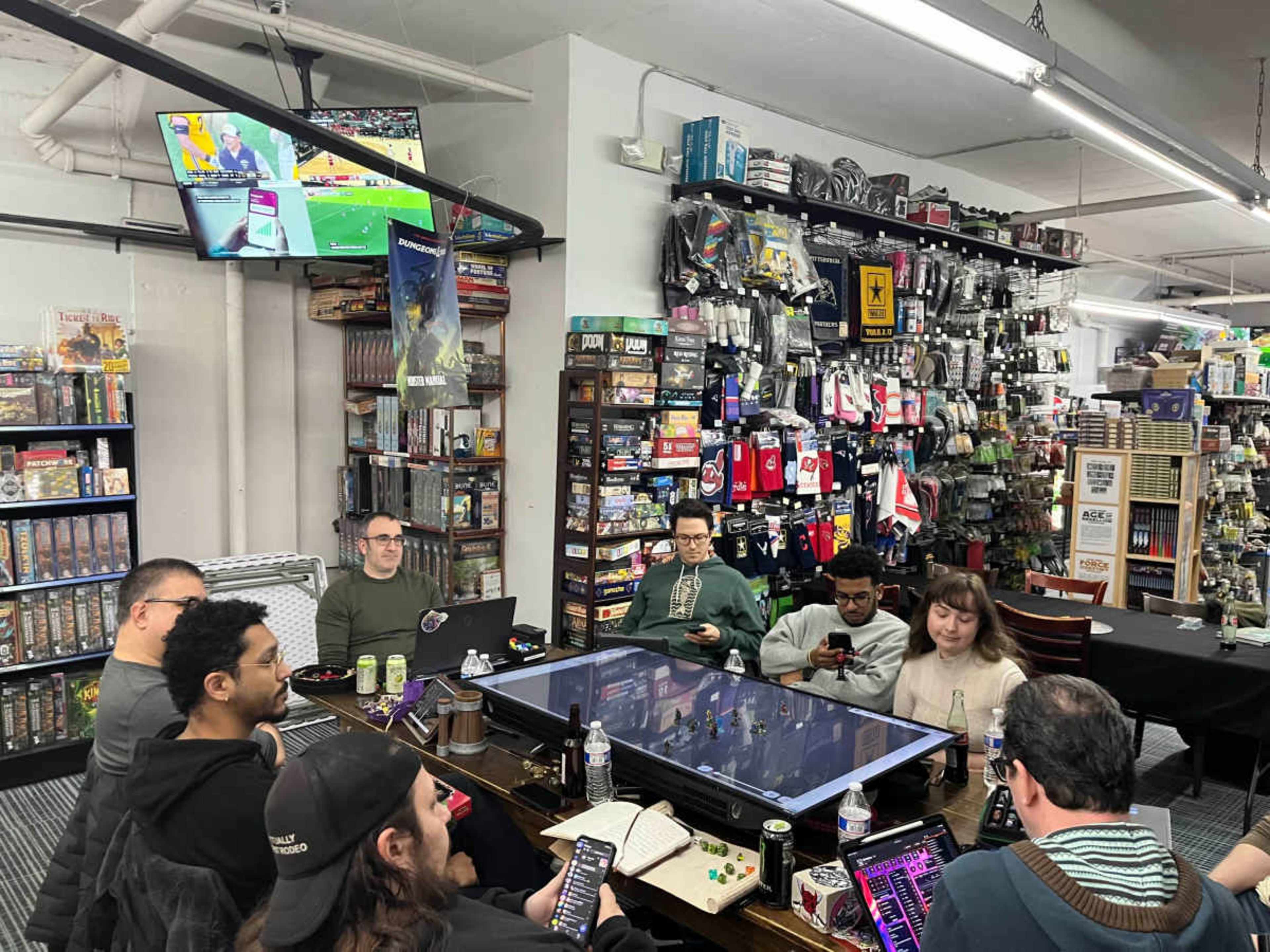 A group of people sits around a large table in a game store, engaging with their devices while various board games and gaming merchandise are displayed on the shelves behind them.
