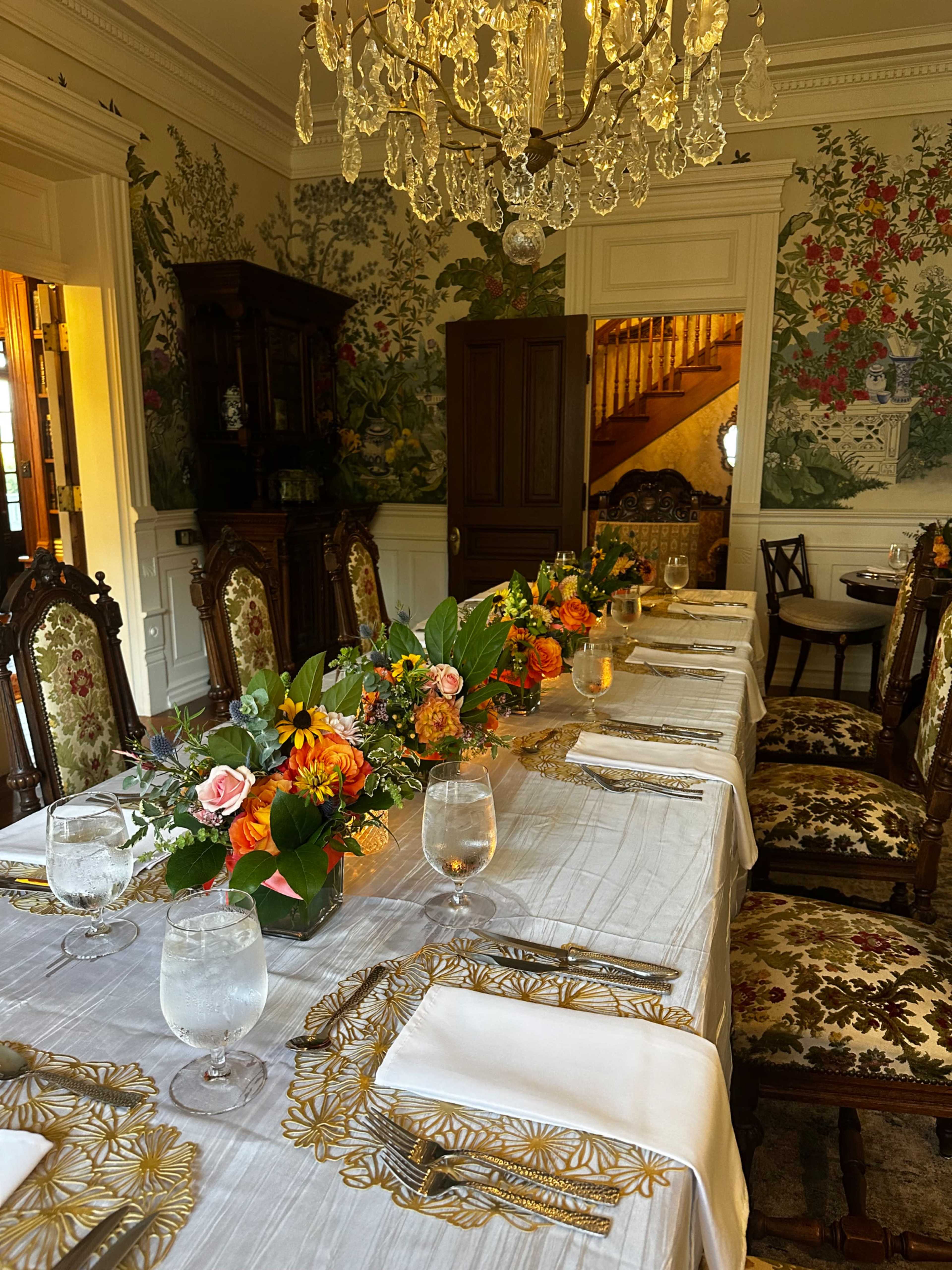 A long dining table is elegantly set with floral arrangements and glasses of water, surrounded by ornate chairs in a room adorned with colorful wallpaper and a chandelier.