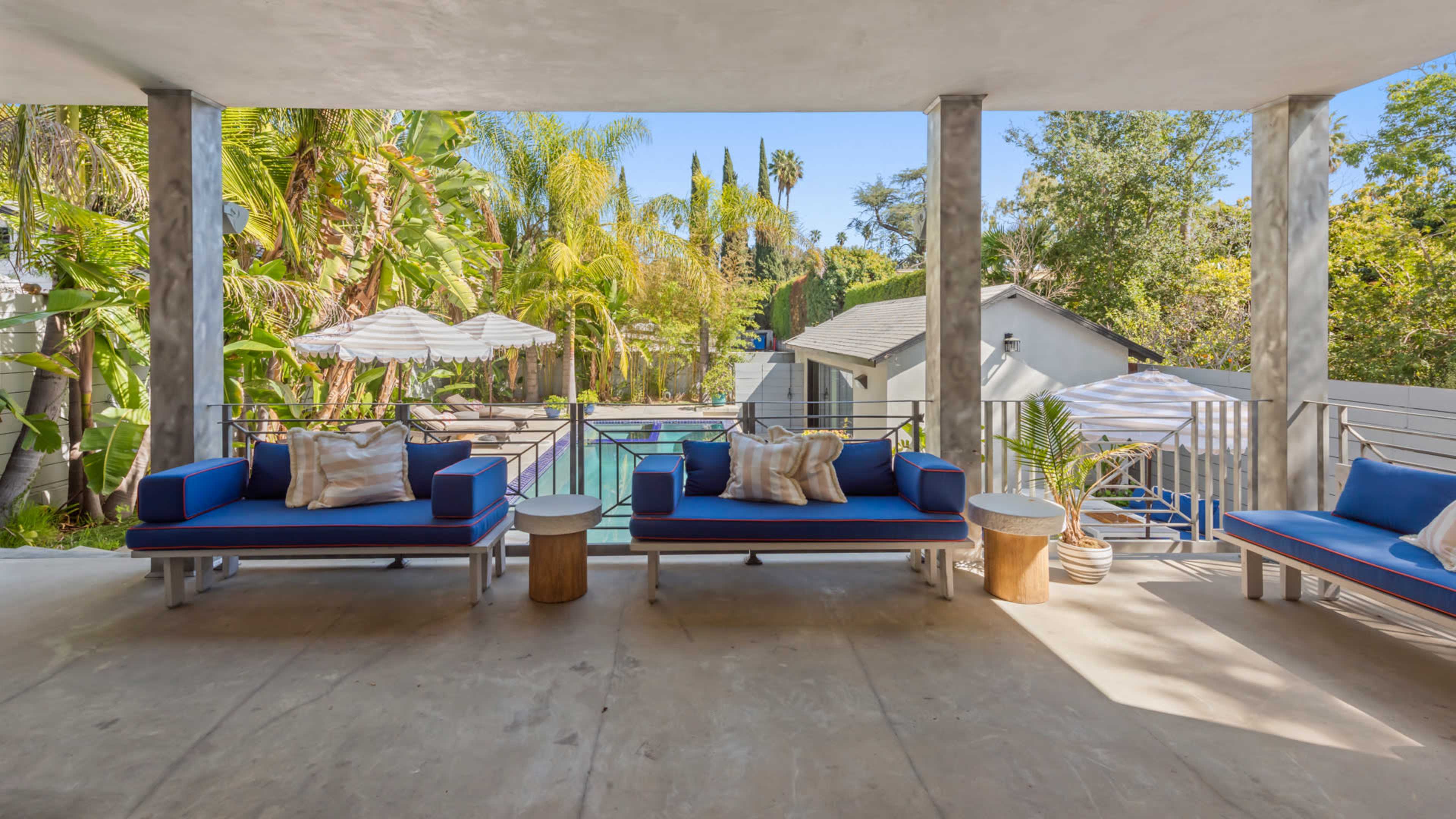 The image shows a patio area with blue couches and a small table overlooking a pool surrounded by tropical plants and trees.
