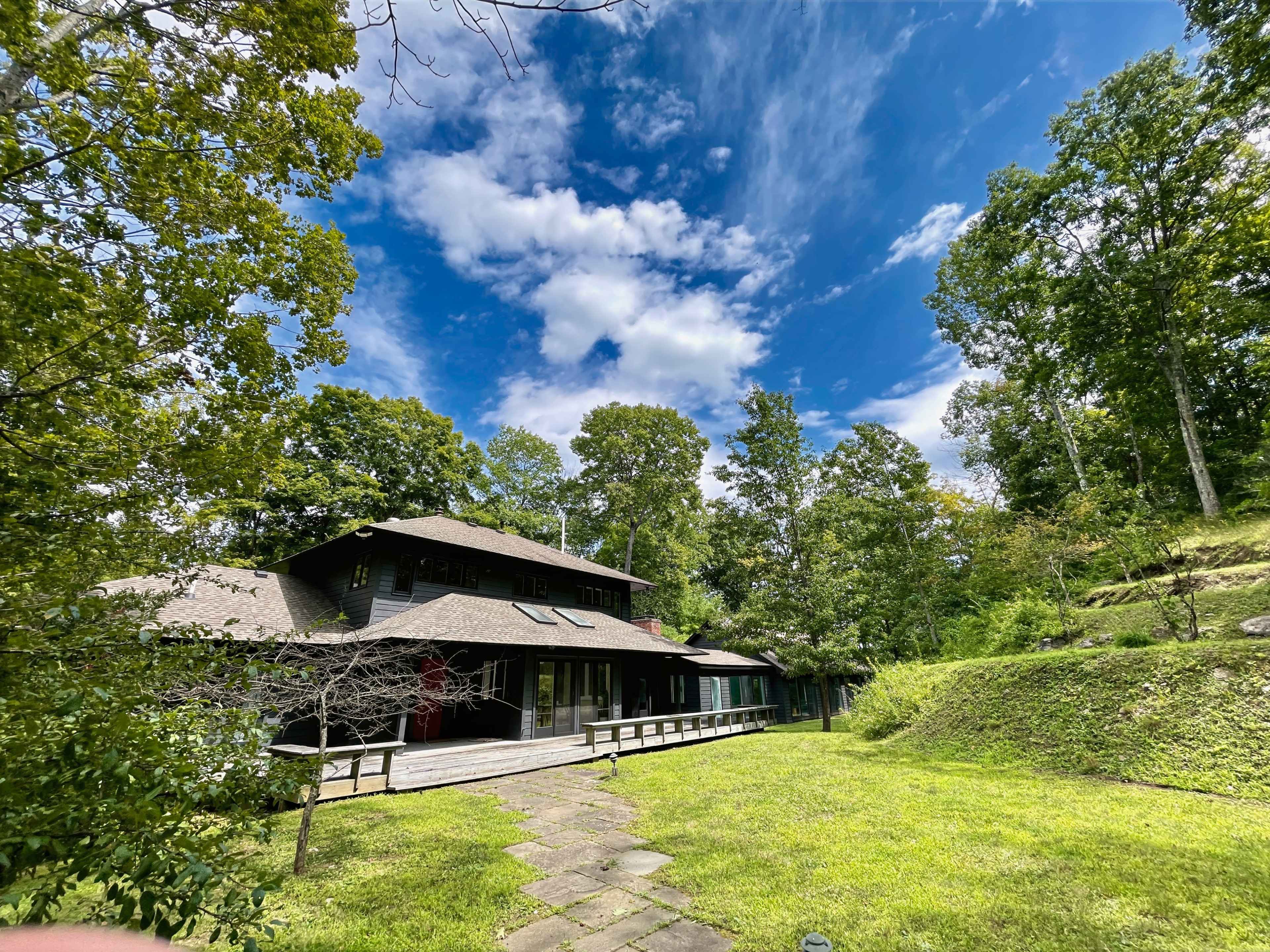 A large house with a gabled roof is set against a backdrop of green trees and a blue sky with scattered clouds.