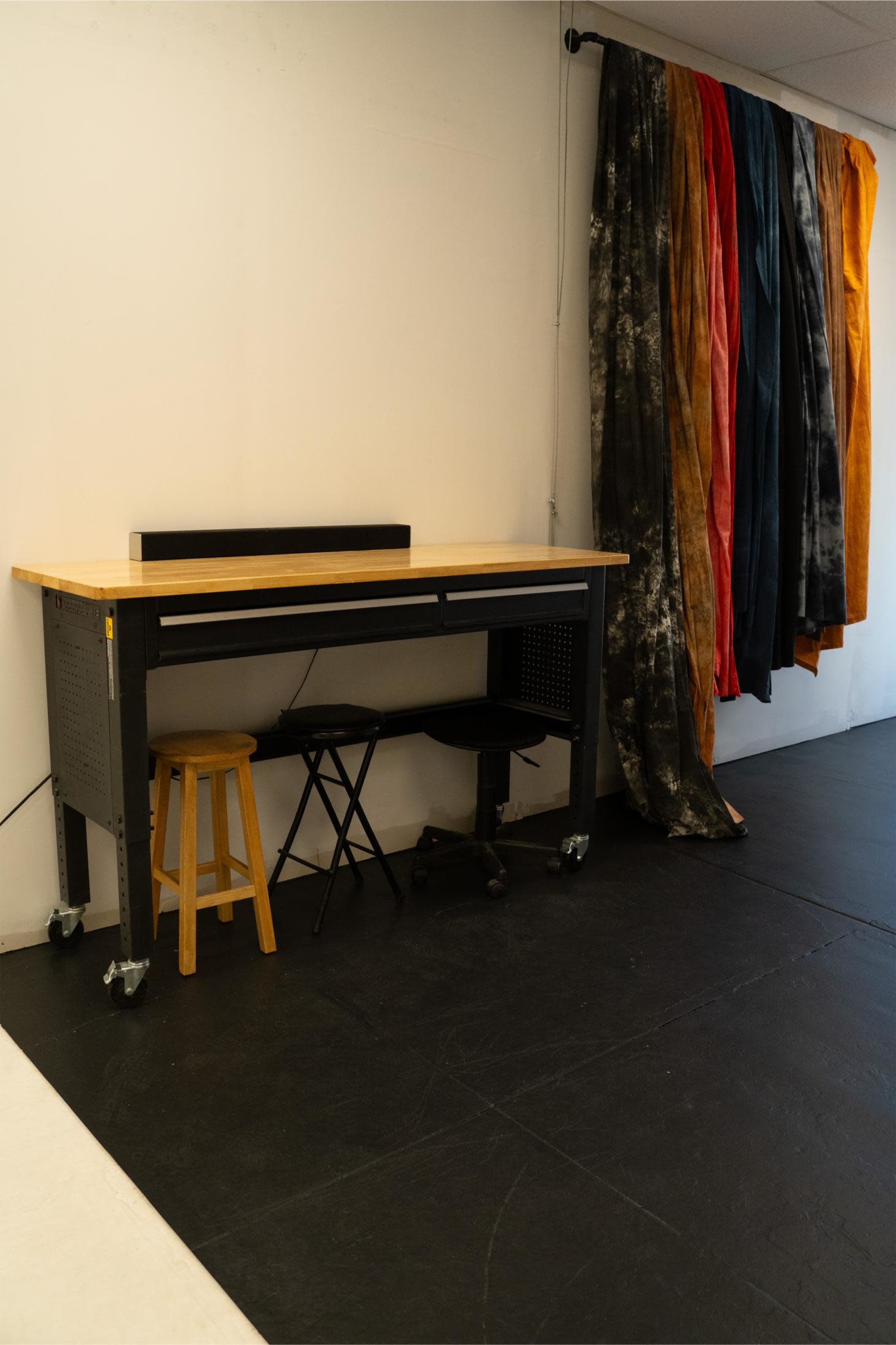 The image shows a workbench with a wooden top and black frame, accompanied by a wooden stool, adjacent to fabric drapes in various colors hanging on the wall.