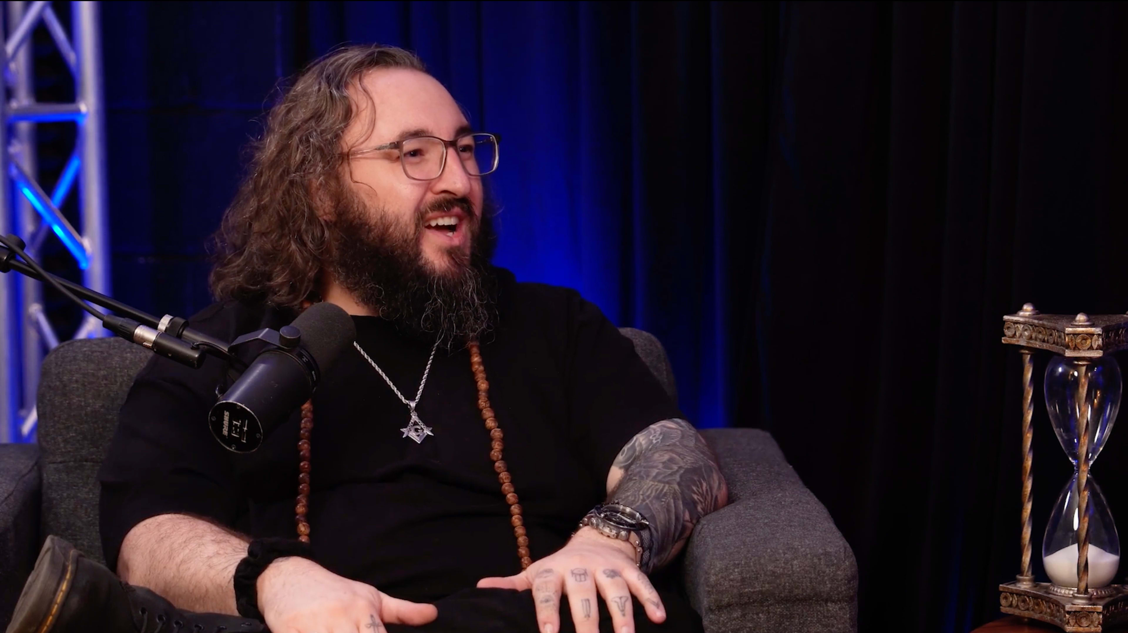 A man with long curly hair and glasses sits on a gray couch, speaking into a microphone.