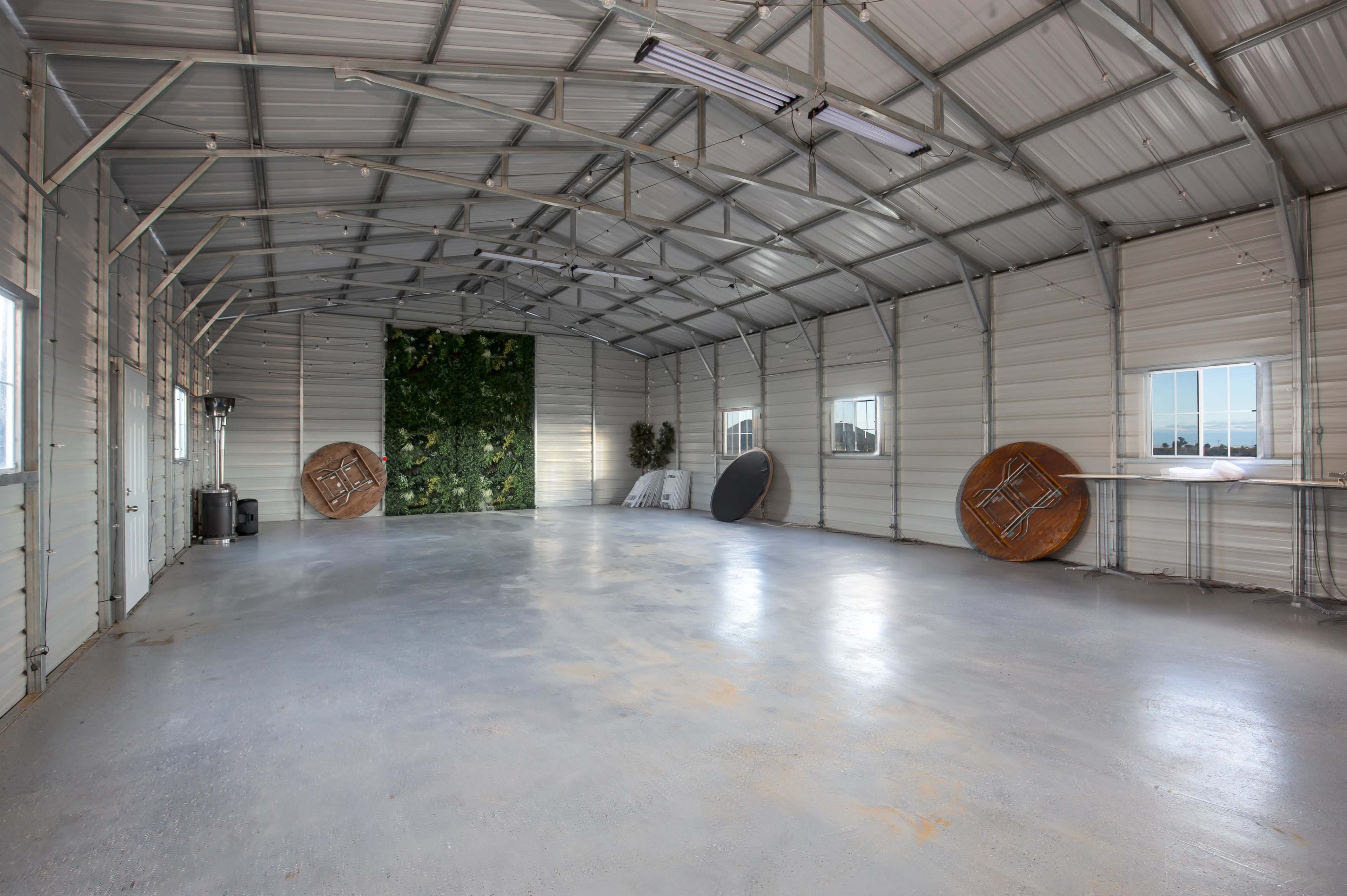 The image shows a spacious, empty metal garage with a concrete floor, featuring large windows and two round tables leaning against the walls.