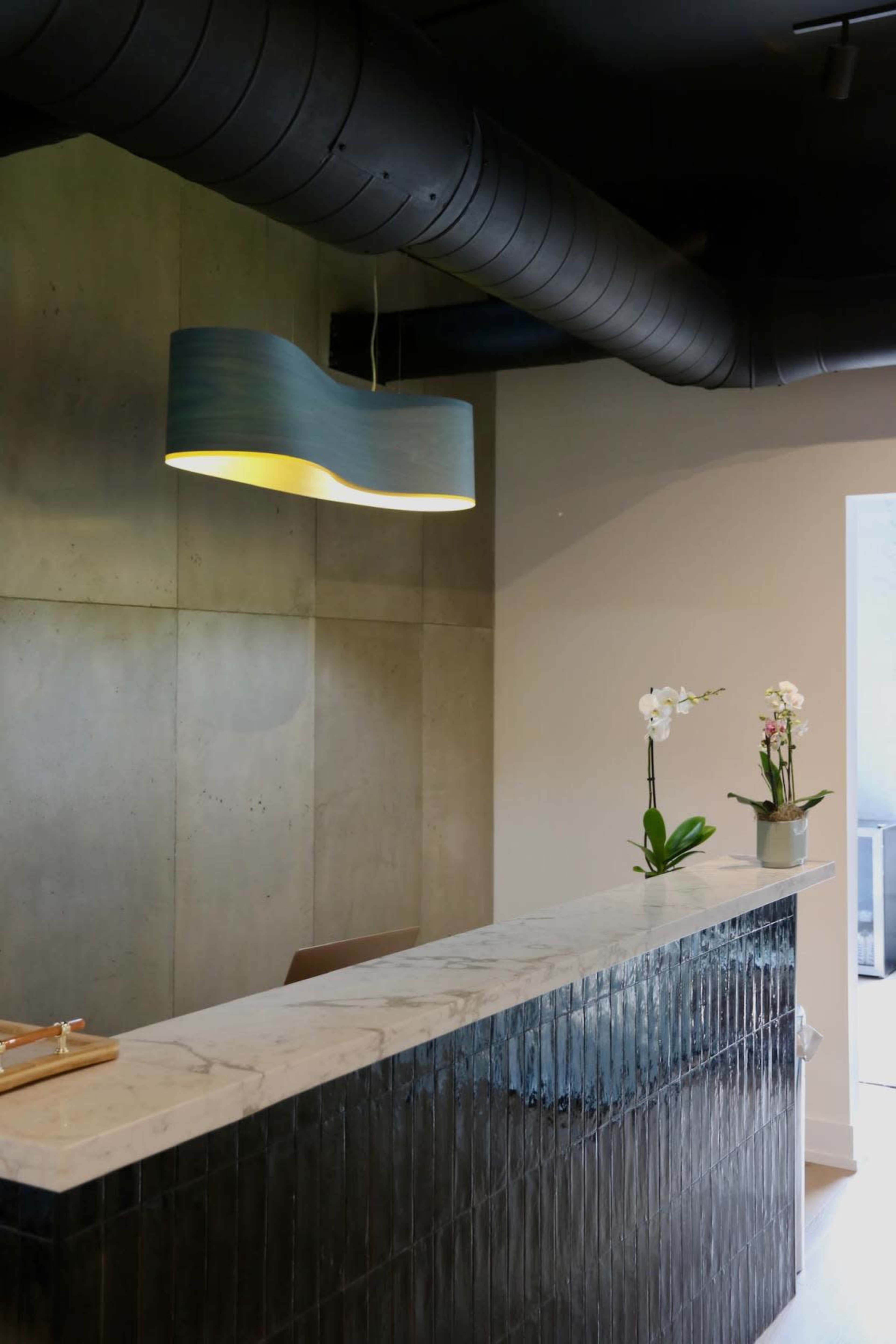 The image shows a modern reception desk made of marble and dark textured panels, with a blue pendant light hanging above and orchid plants placed on the counter.