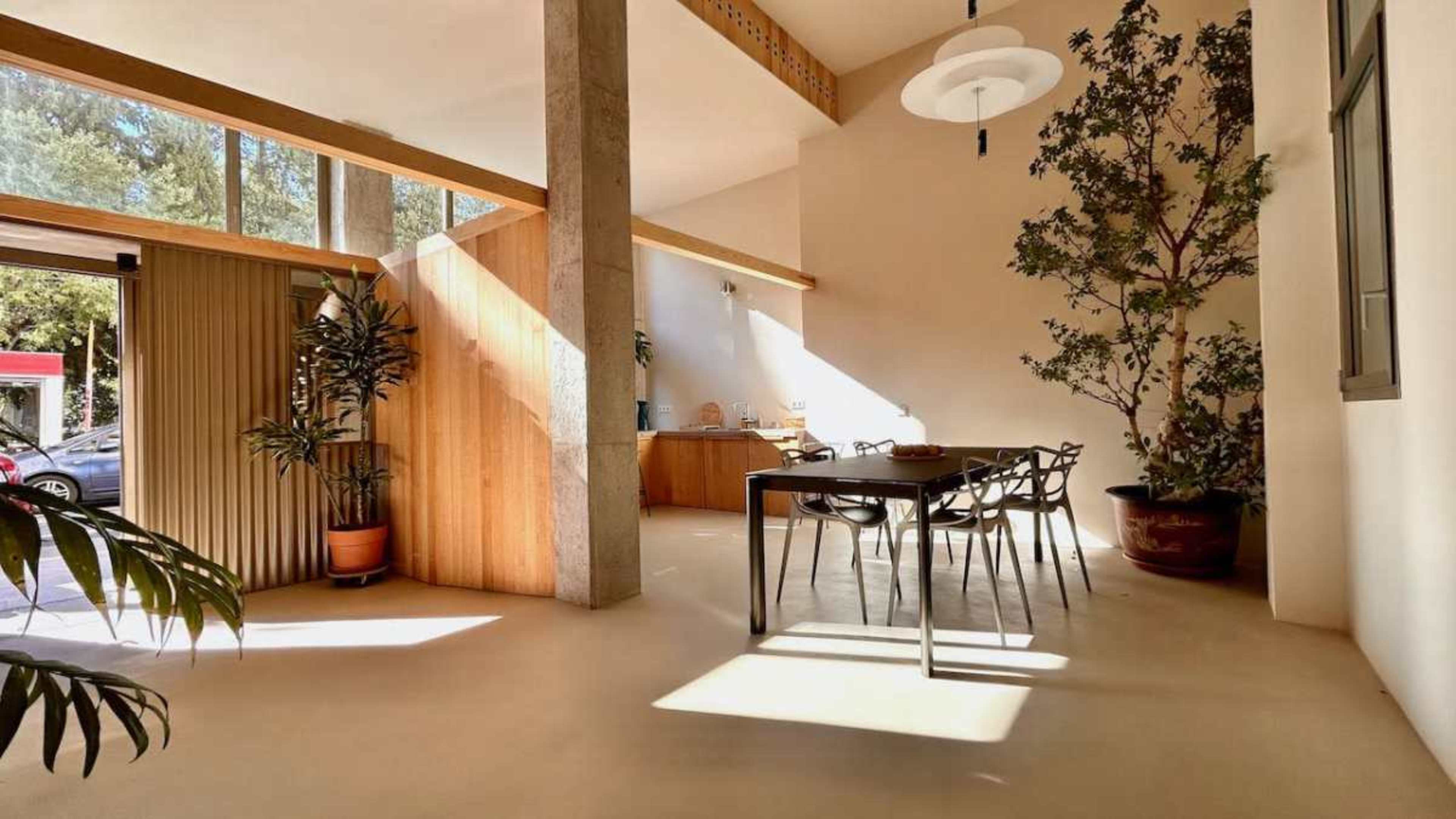 A bright dining area features a wooden table surrounded by chairs, with large windows allowing natural light to illuminate the space and a potted plant in the corner.