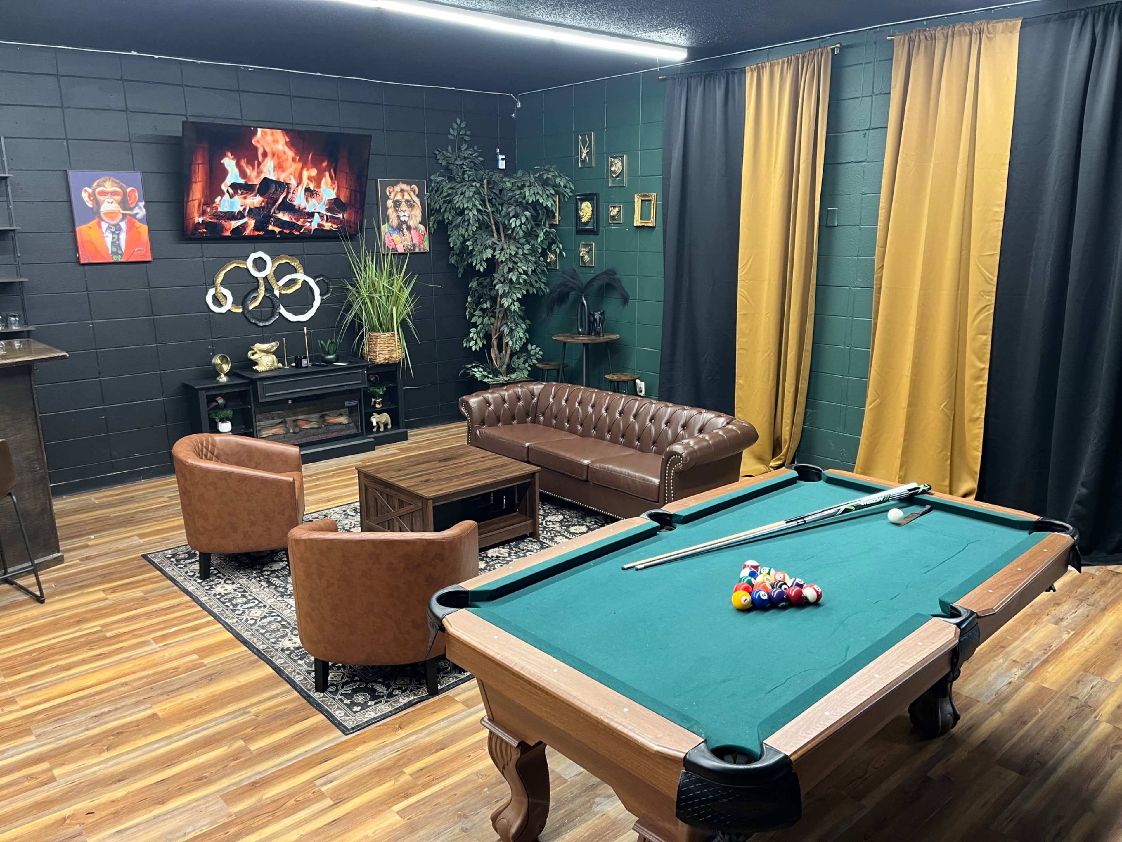 The image shows a stylish game room featuring a pool table, brown leather chairs, a sofa, and a decorative wall with a faux fireplace and artwork.