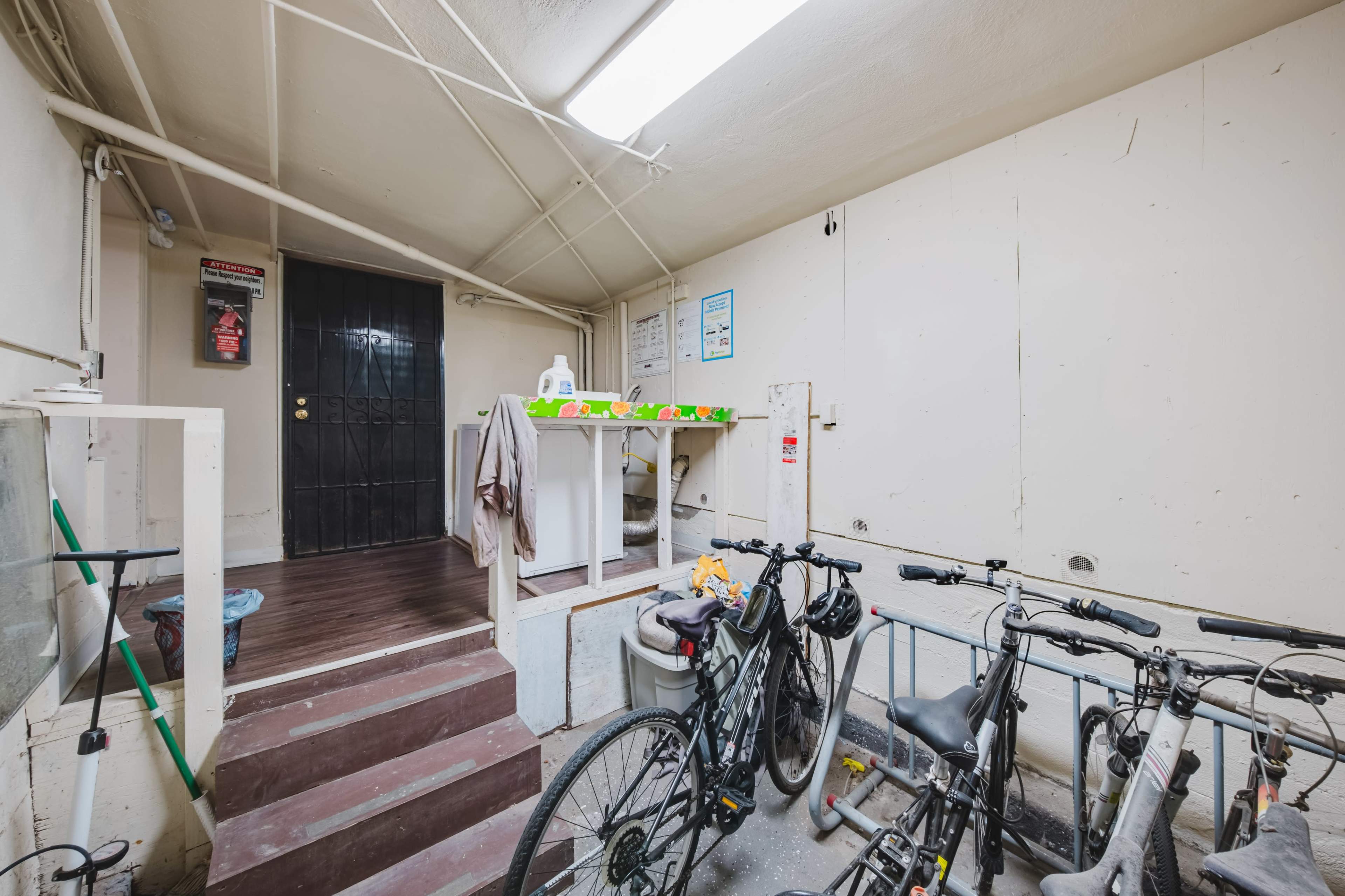 The image shows a dimly lit hallway with a stairway leading to a door, bicycles parked on the floor, and a table with items atop it.