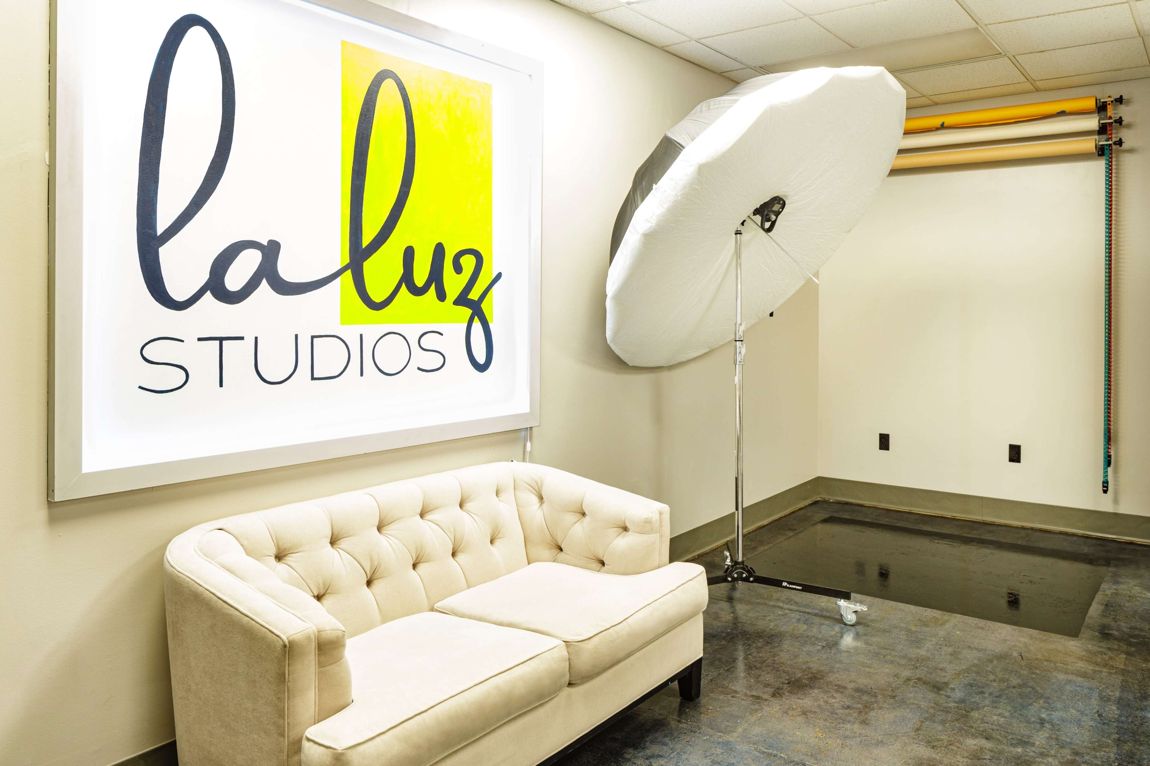 A well-lit studio space featuring a tufted beige sofa, a large backlit sign reading "la luz STUDIOS," and a photography umbrella on a stand next to a backdrop.