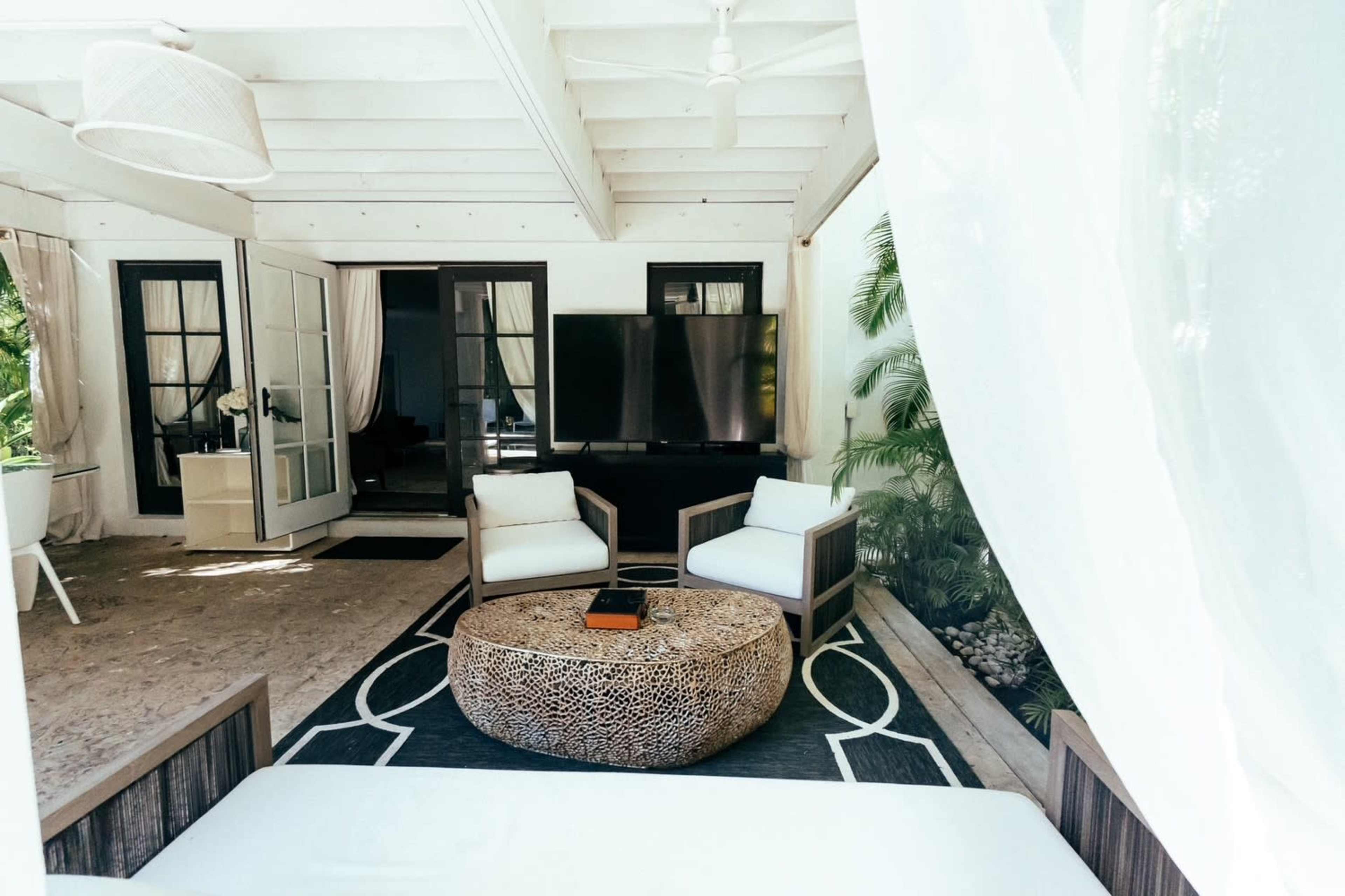 The image shows a cozy outdoor seating area with two chairs, a round coffee table, and a large television, surrounded by lush greenery.